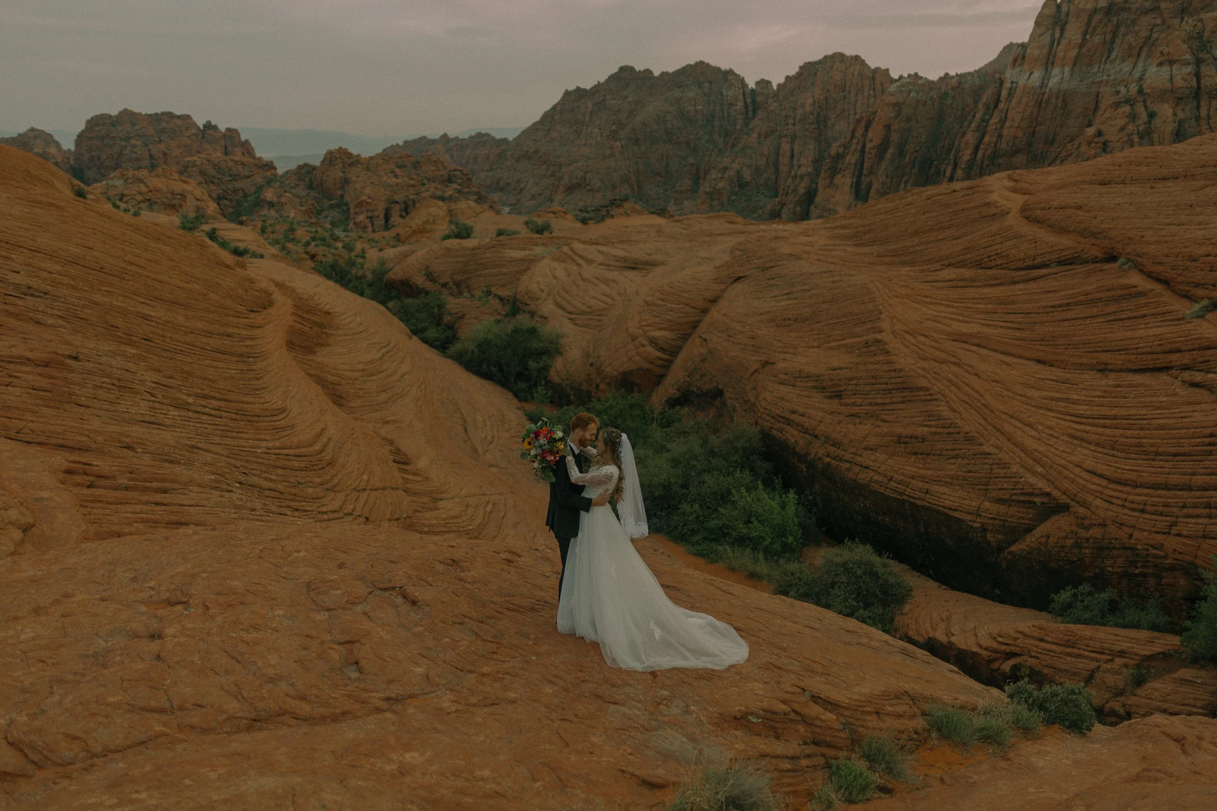 Bride and groom kissing in a desert landscape with red rock formations and greenery.