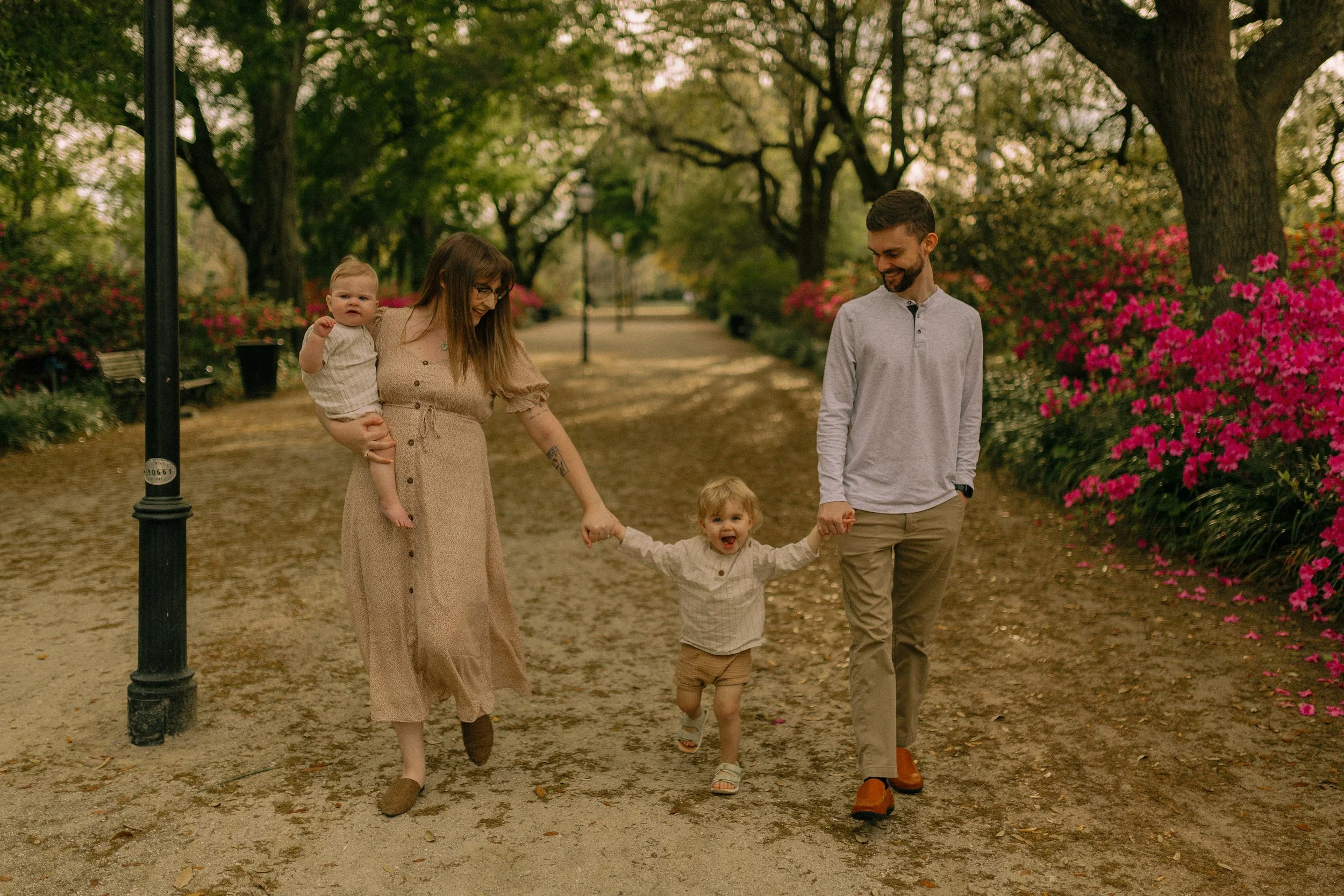 Family walking in a park holding hands, with two children, surrounded by trees and pink flowers.