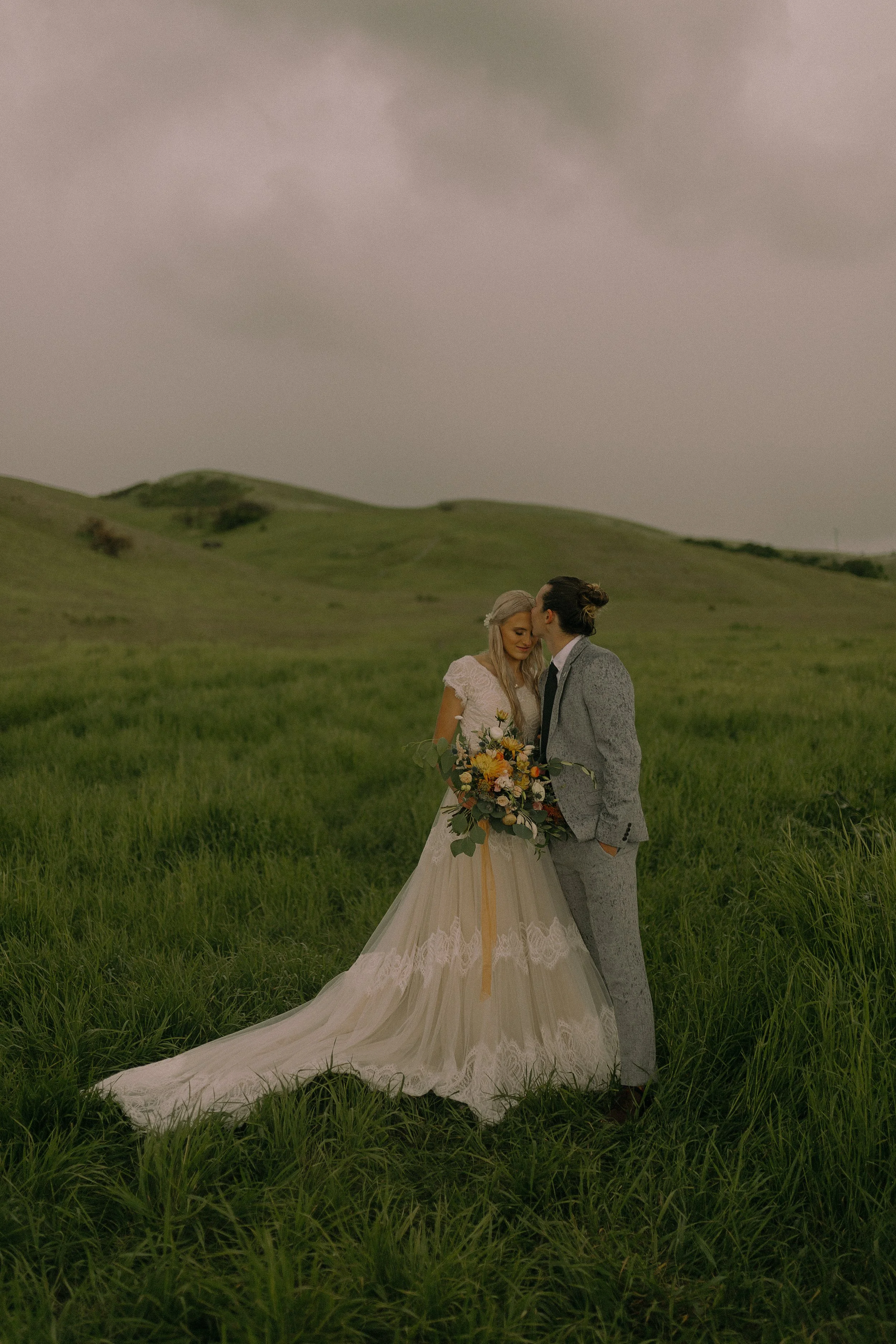 Bride and groom standing in a grassy field with the groom kissing the bride's temple. The bride holds a bouquet of flowers, and both are dressed in formal wedding attire.