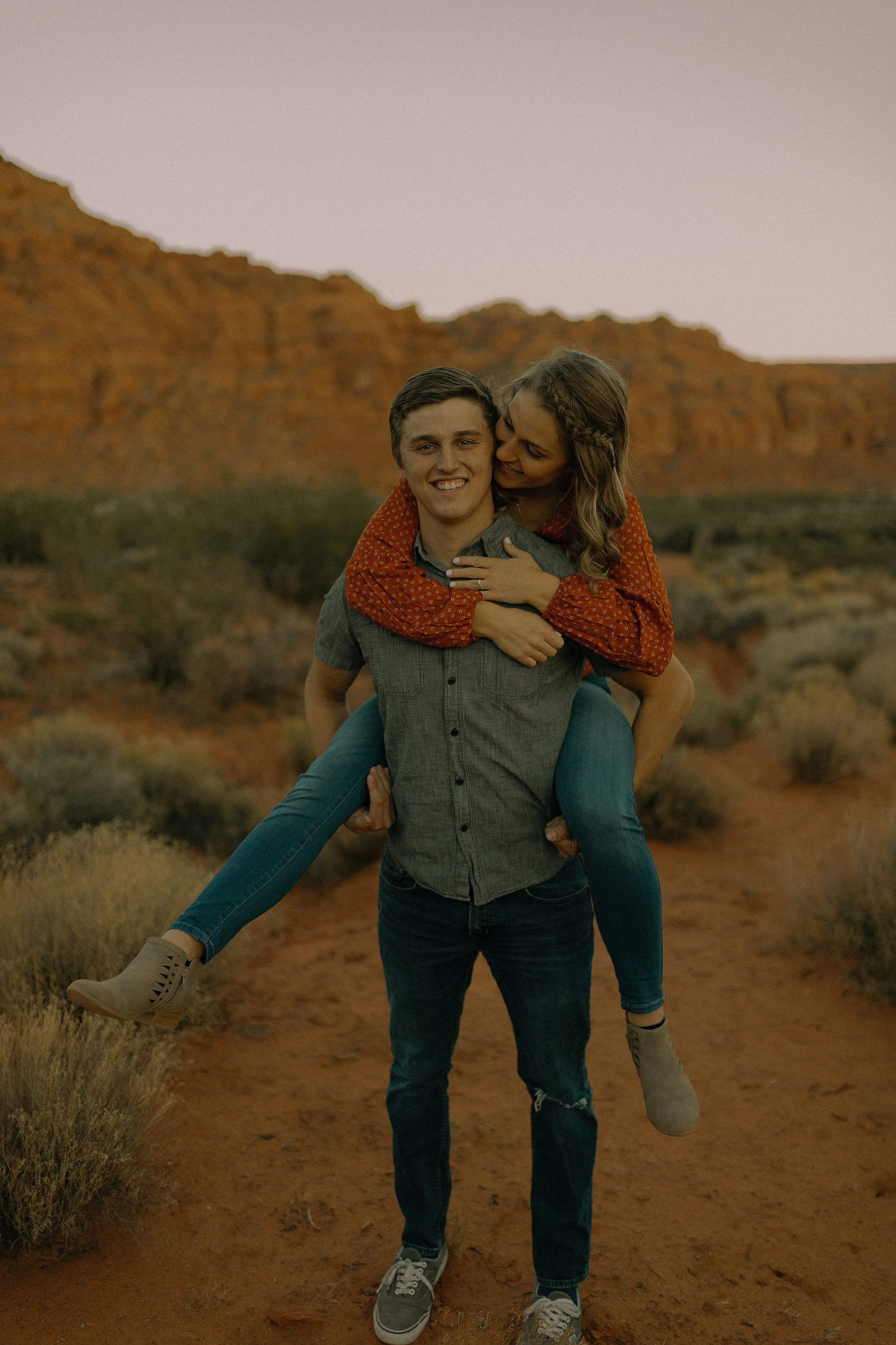 A couple in a desert landscape, with a woman in a red top and jeans playfully riding on a man's back. He is wearing a gray shirt and jeans. The background features red rock formations and desert vegetation.