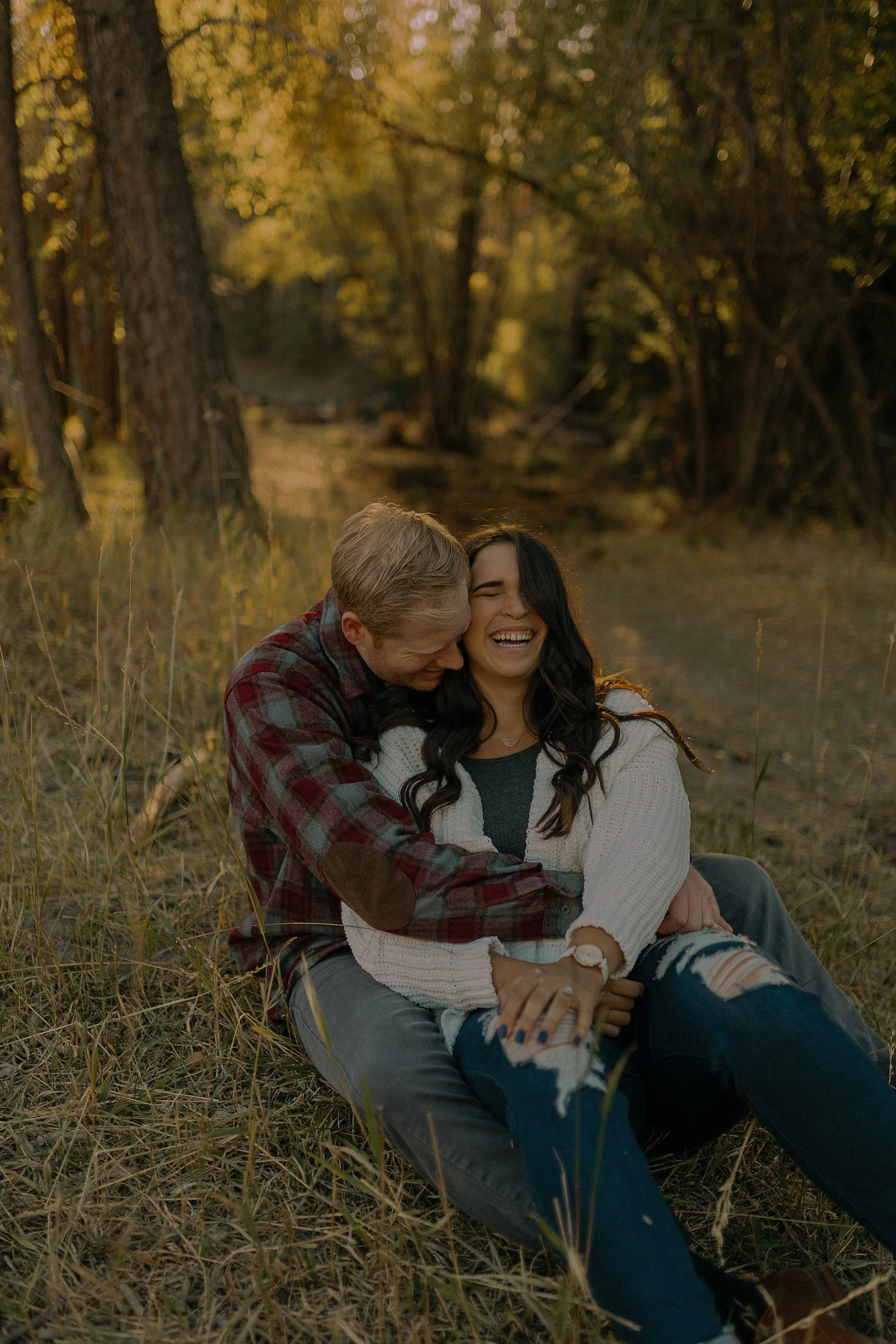 Couple sitting in a forest, woman wearing a white sweater and jeans, man in a plaid shirt, both laughing and embracing.