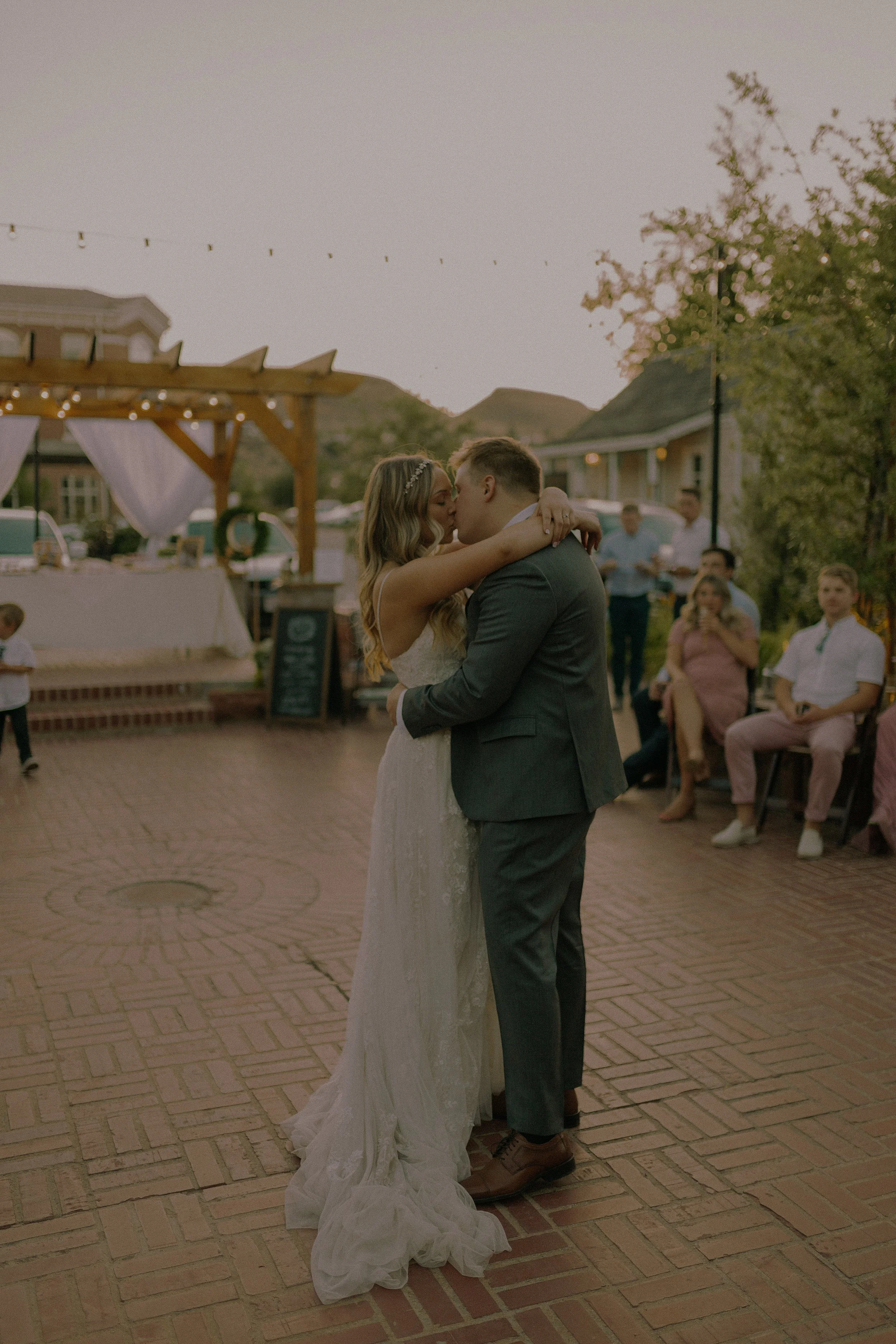 A bride and groom embrace during their first dance at an outdoor wedding reception. Guests are seated, watching the couple. The setting features a wooden pergola with string lights and nearby greenery.