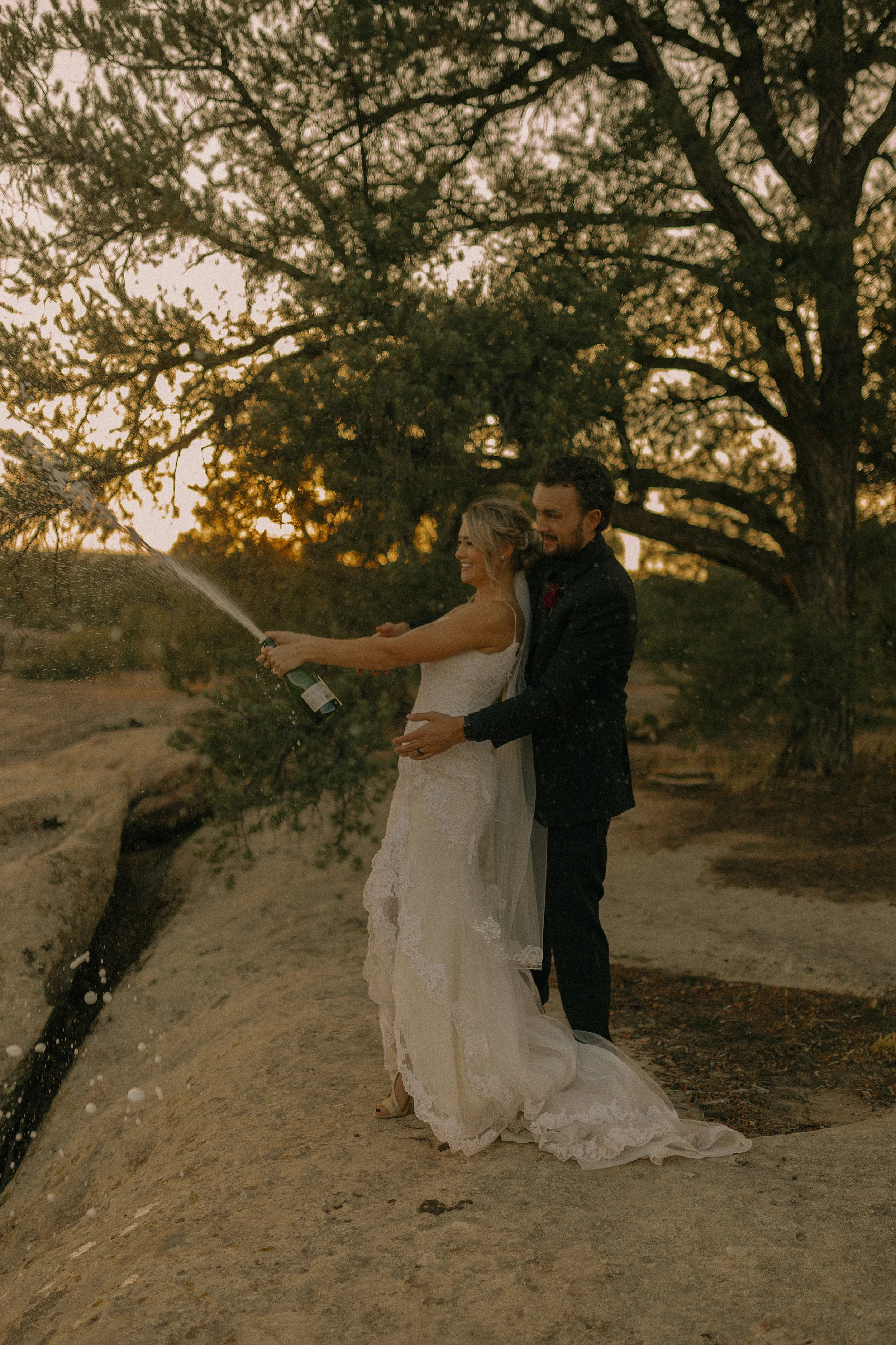 Bride and groom celebrating outdoors, popping a champagne bottle with trees and sunset in the background.