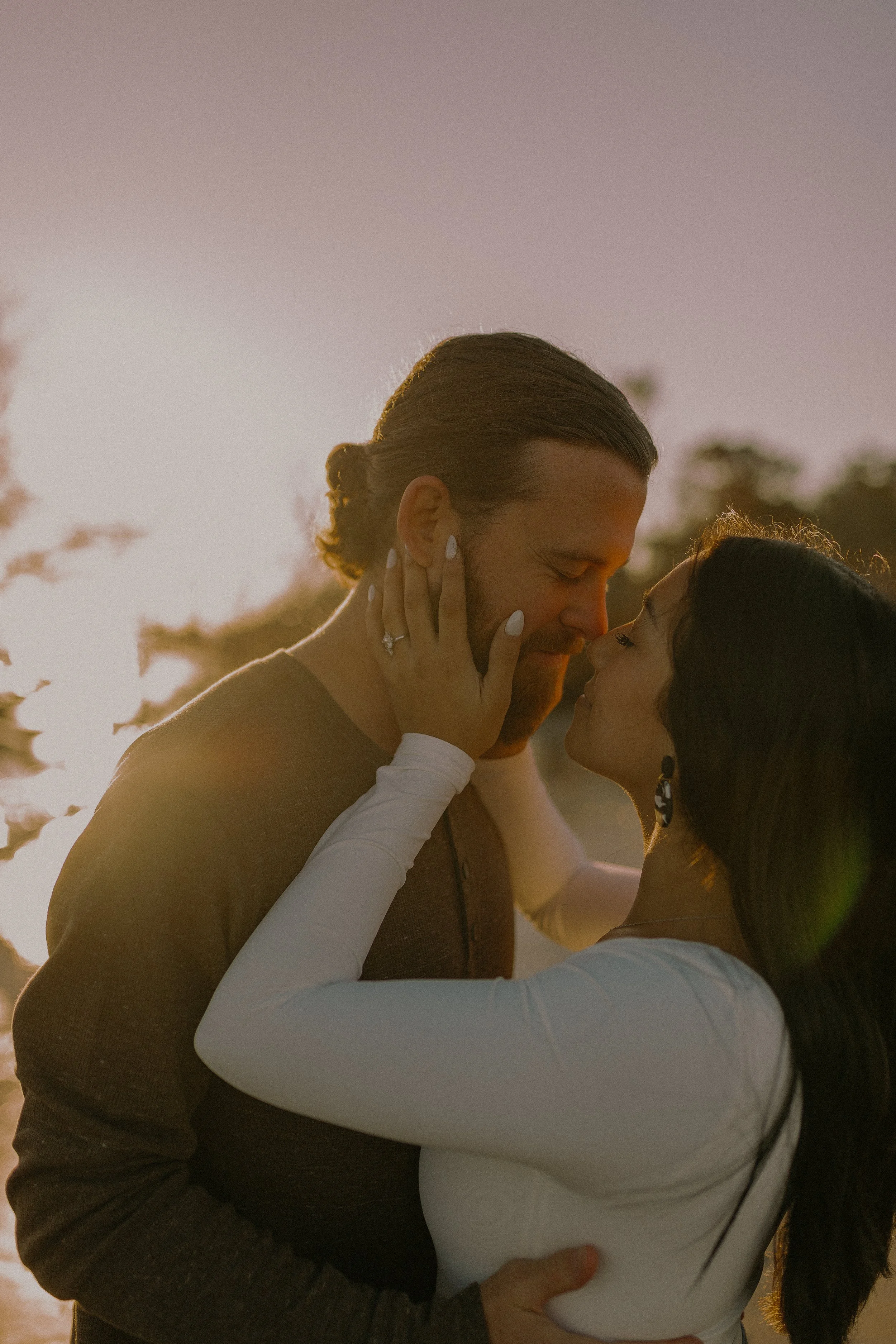 A couple sharing an intimate moment at sunset, with soft lighting and a natural background. One person's hand is on the other's cheek, showcasing a ring.