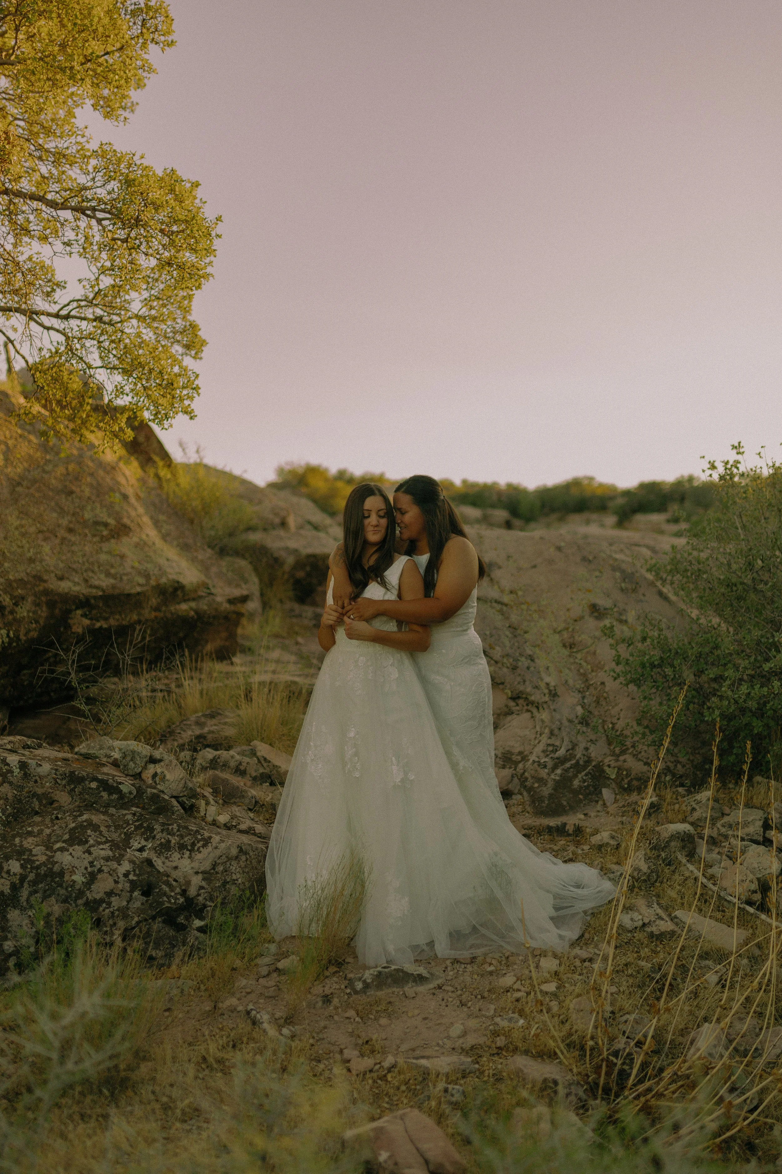Two women in wedding dresses embracing in a rocky outdoor setting during sunset.