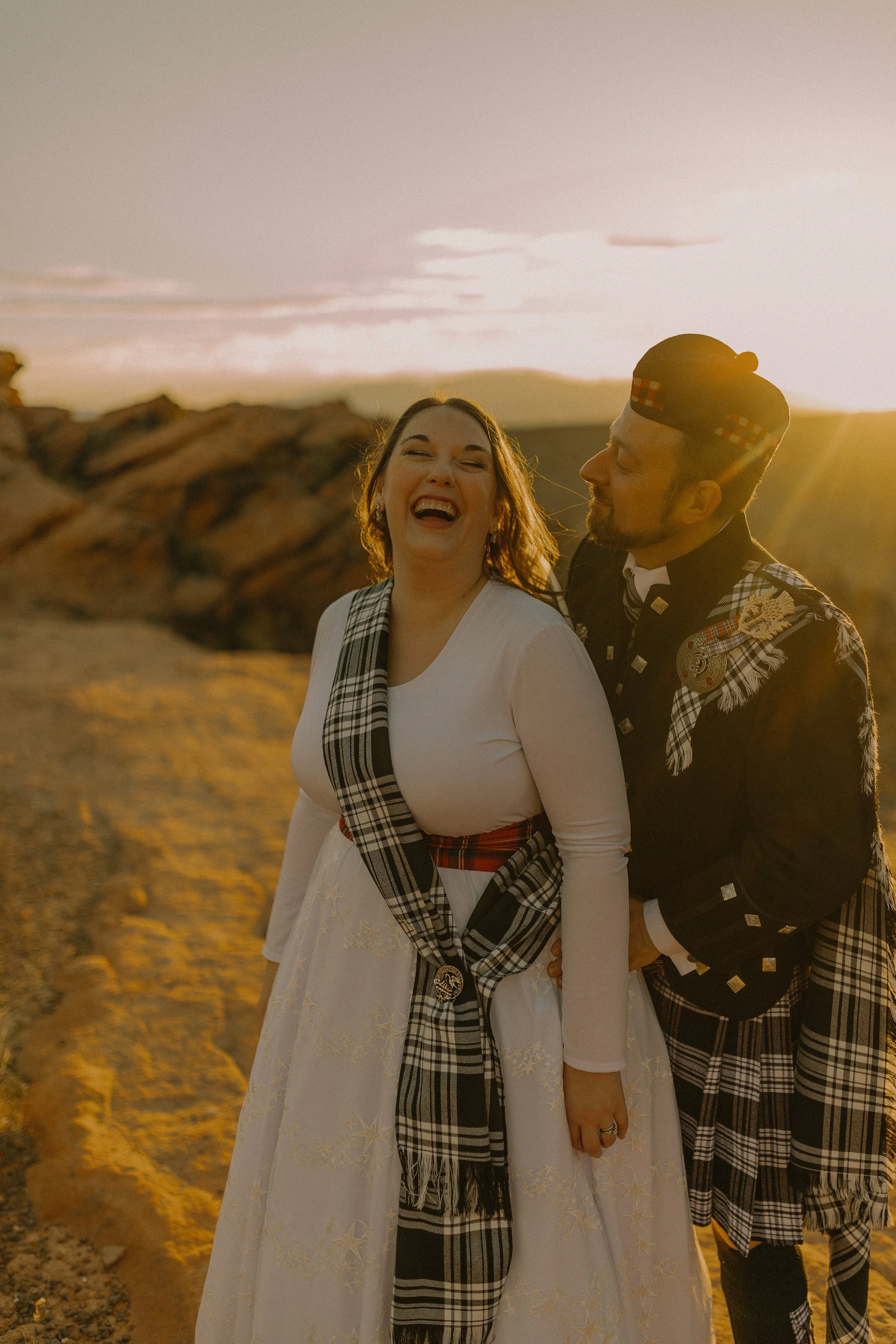 A couple wearing traditional Scottish attire, including kilts and tartan accessories, smiling and embracing at sunset in an outdoor setting.