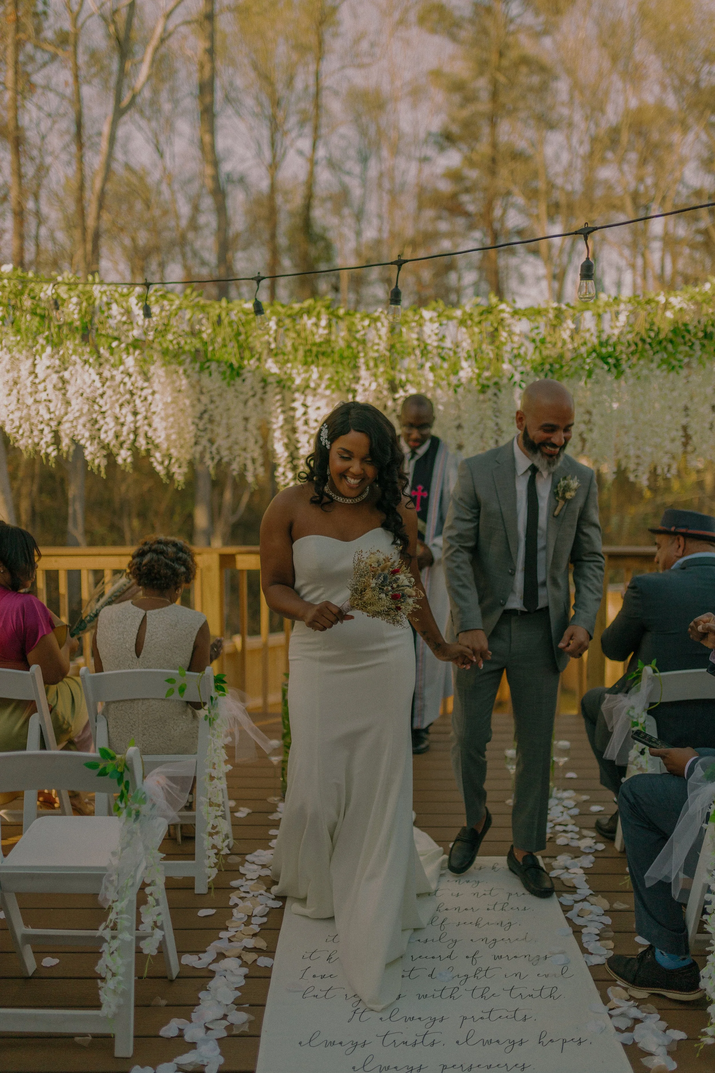 Bride and groom walking down an outdoor wedding aisle, decorated with hanging flowers and string lights, guests seated on either side.