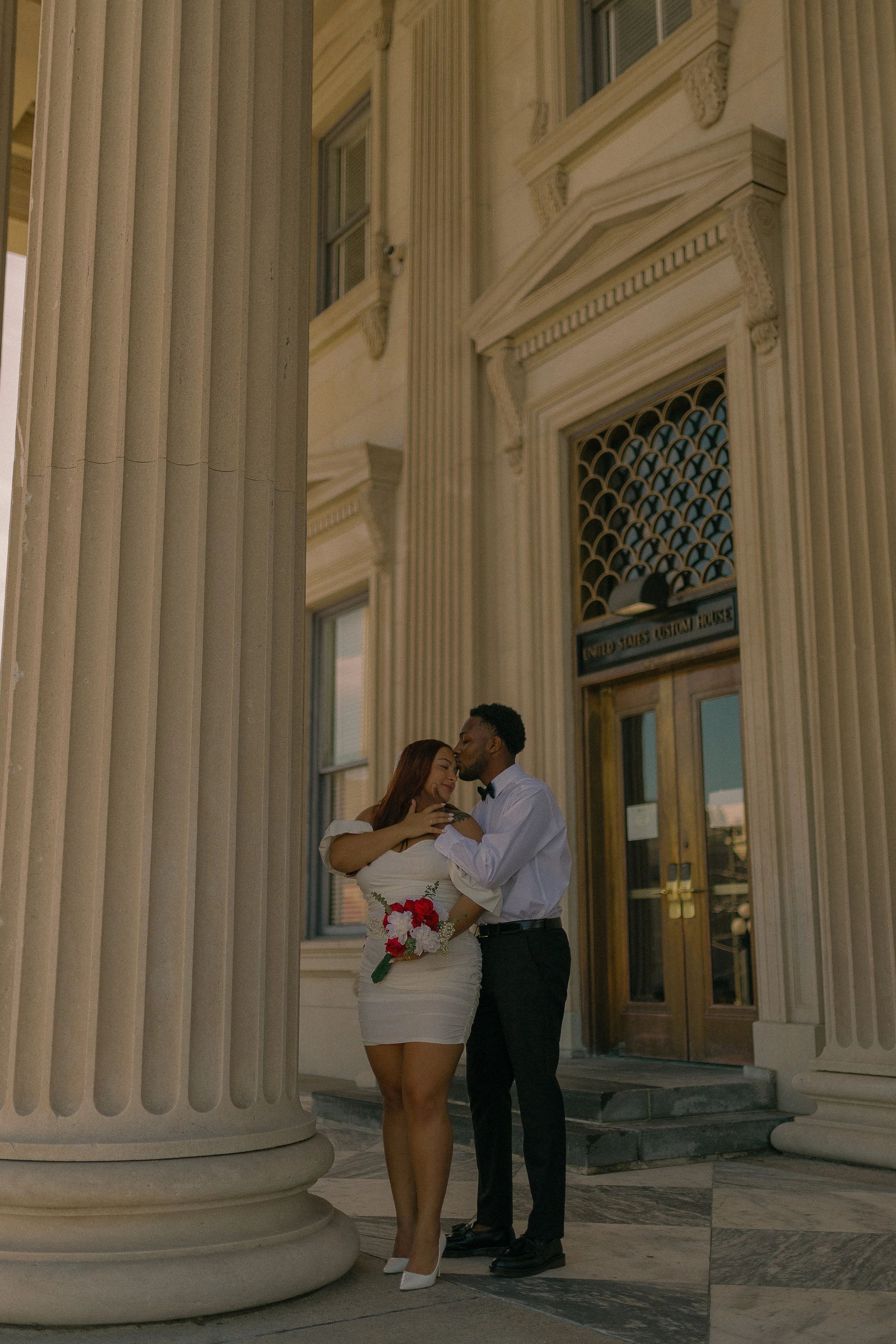 Couple embracing in front of a historic building entrance with Corinthian columns, holding a bouquet.