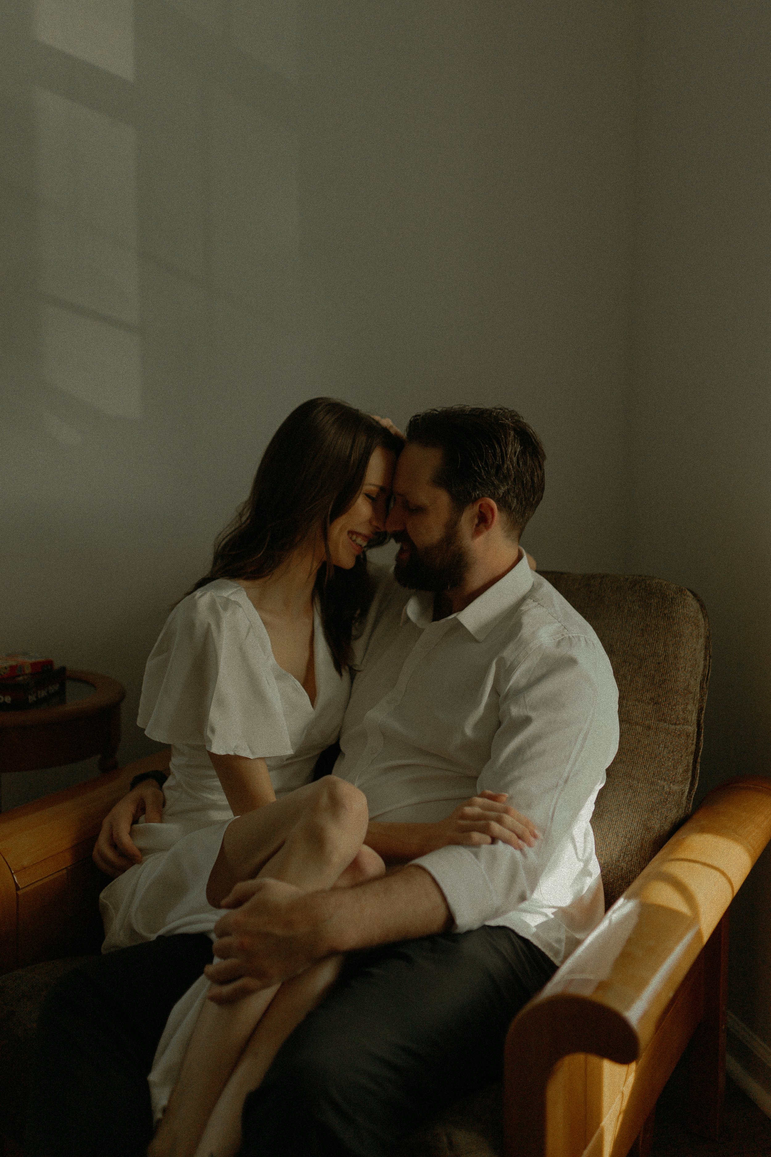 A couple sitting closely together on an armchair, smiling and leaning into each other in a softly lit room.
