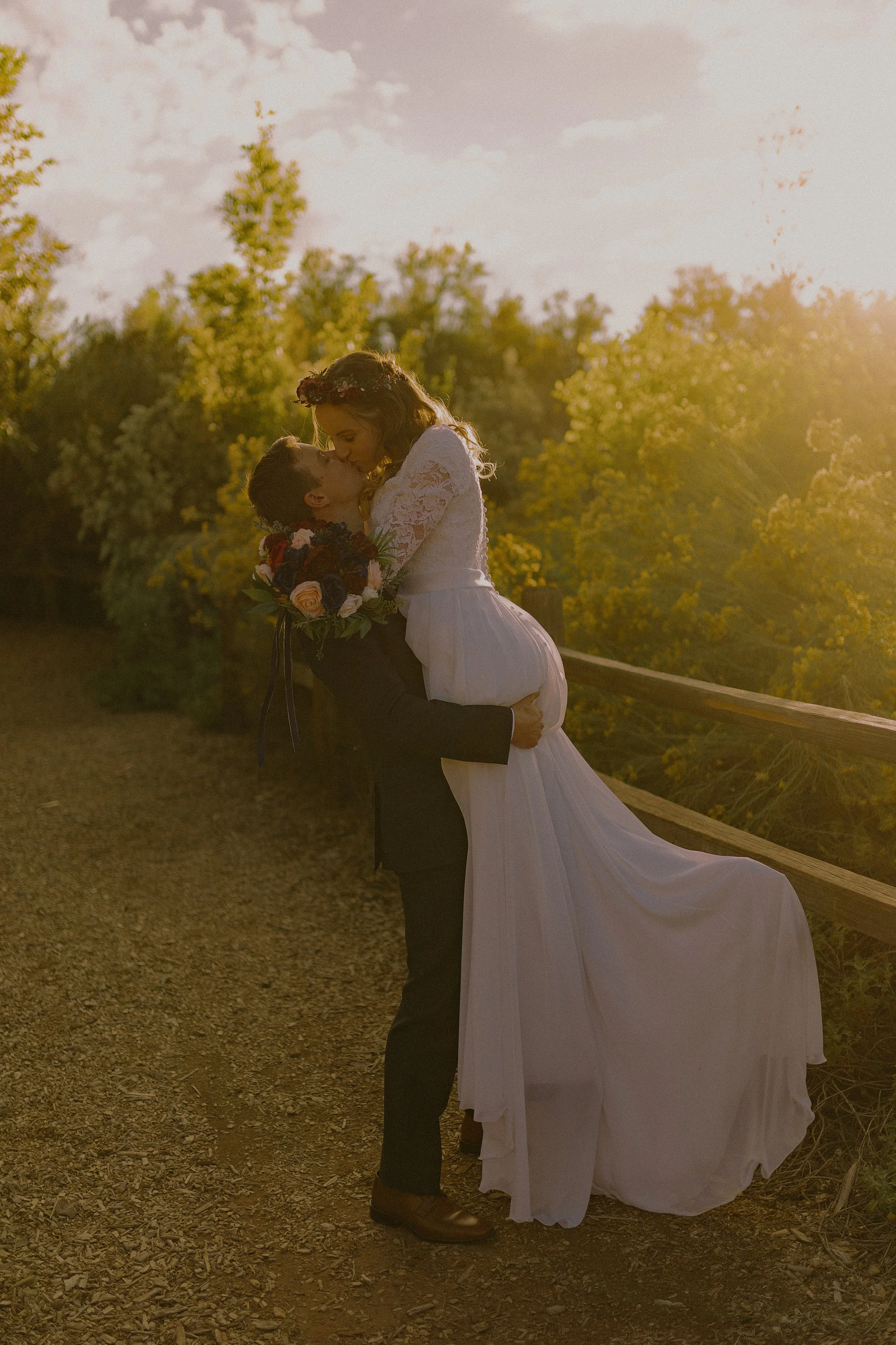Bride and groom kissing outdoors, with the groom lifting the bride. She wears a white dress and holds a bouquet. Sunlight filters through surrounding trees.