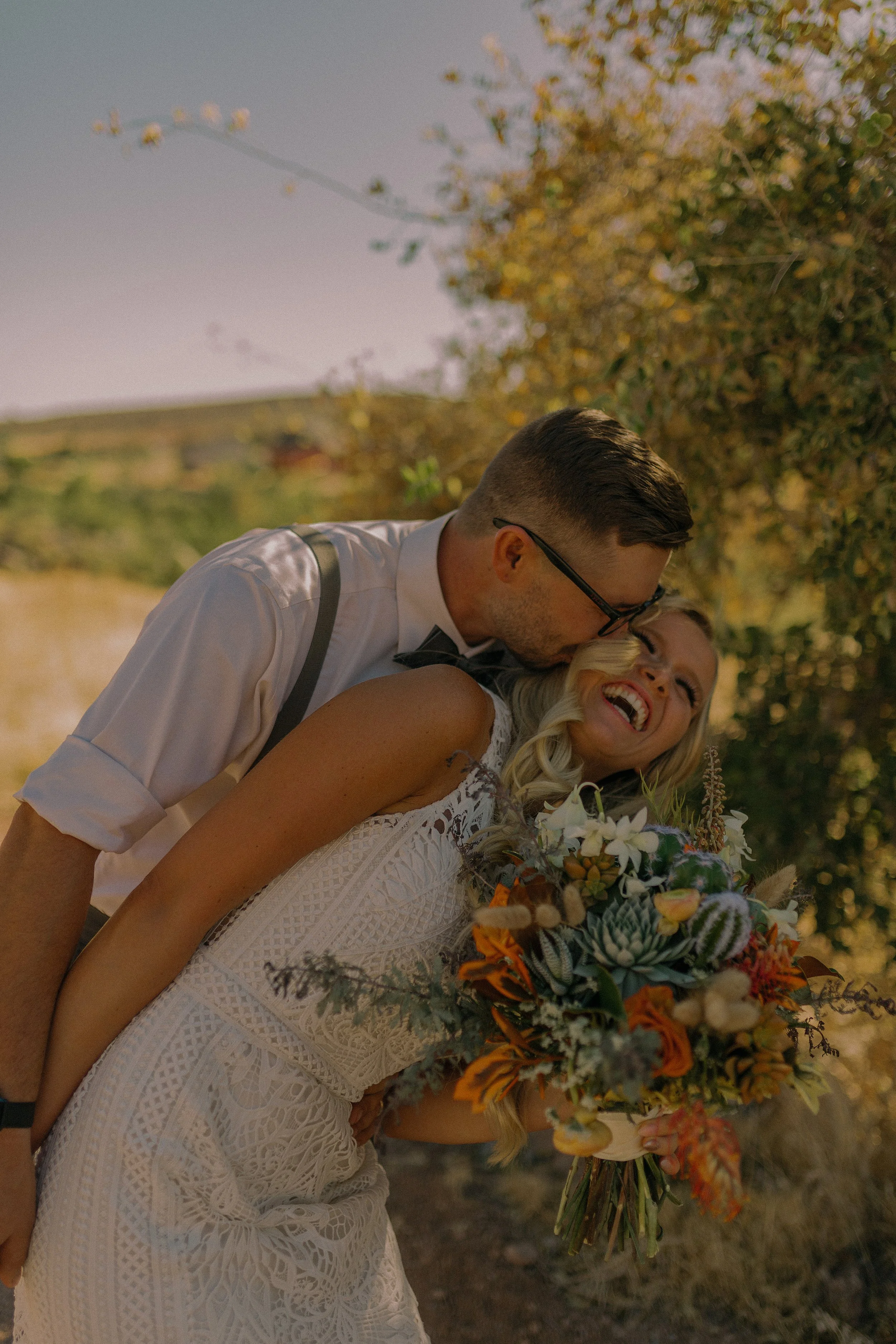 A couple, possibly newlyweds, are embracing outdoors. The woman is holding a colorful bouquet and smiling joyfully while the man kisses her cheek.