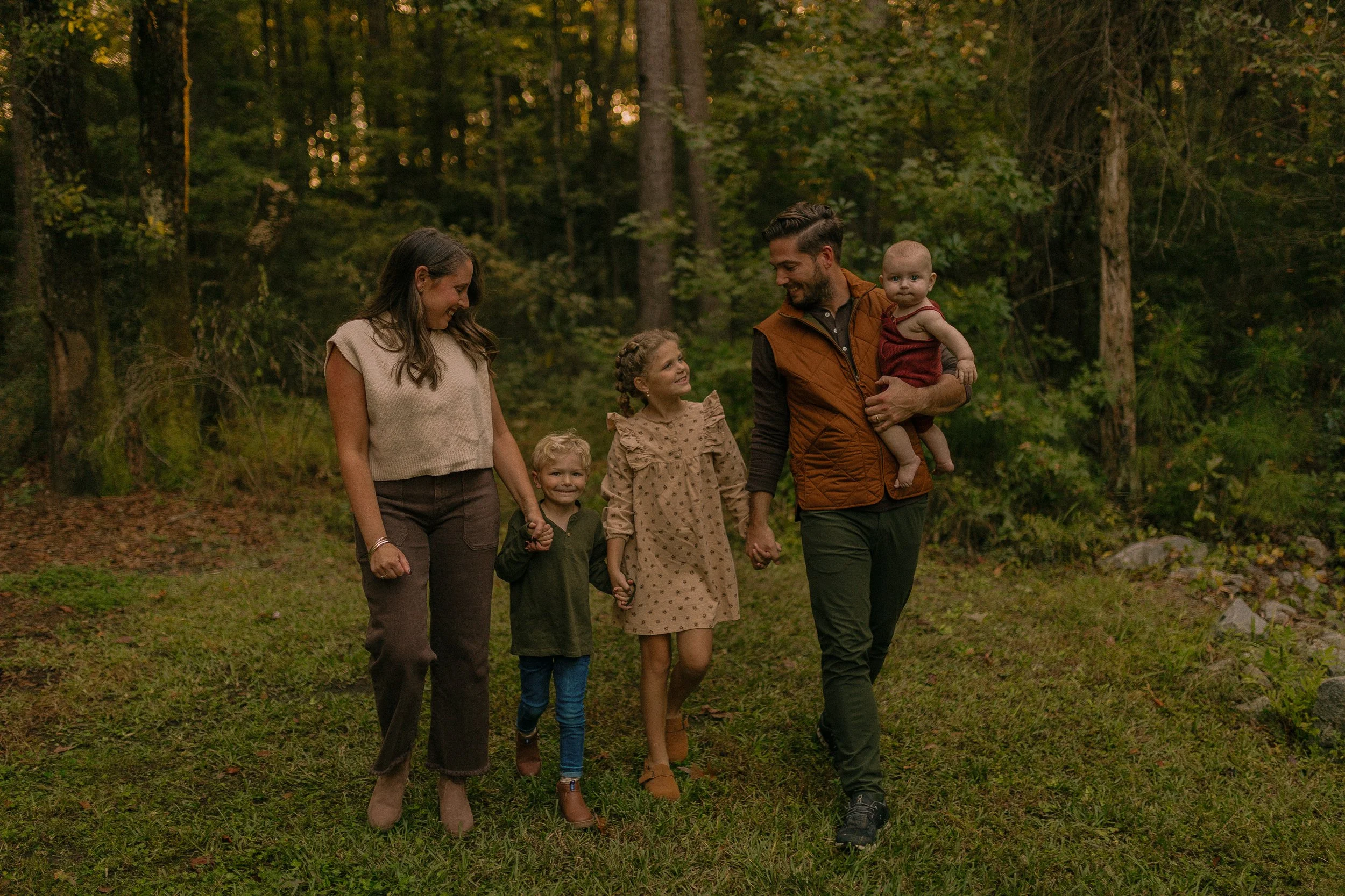 Family walking together in a wooded area, consisting of a man holding a baby, a woman, and two children holding hands, all smiling.