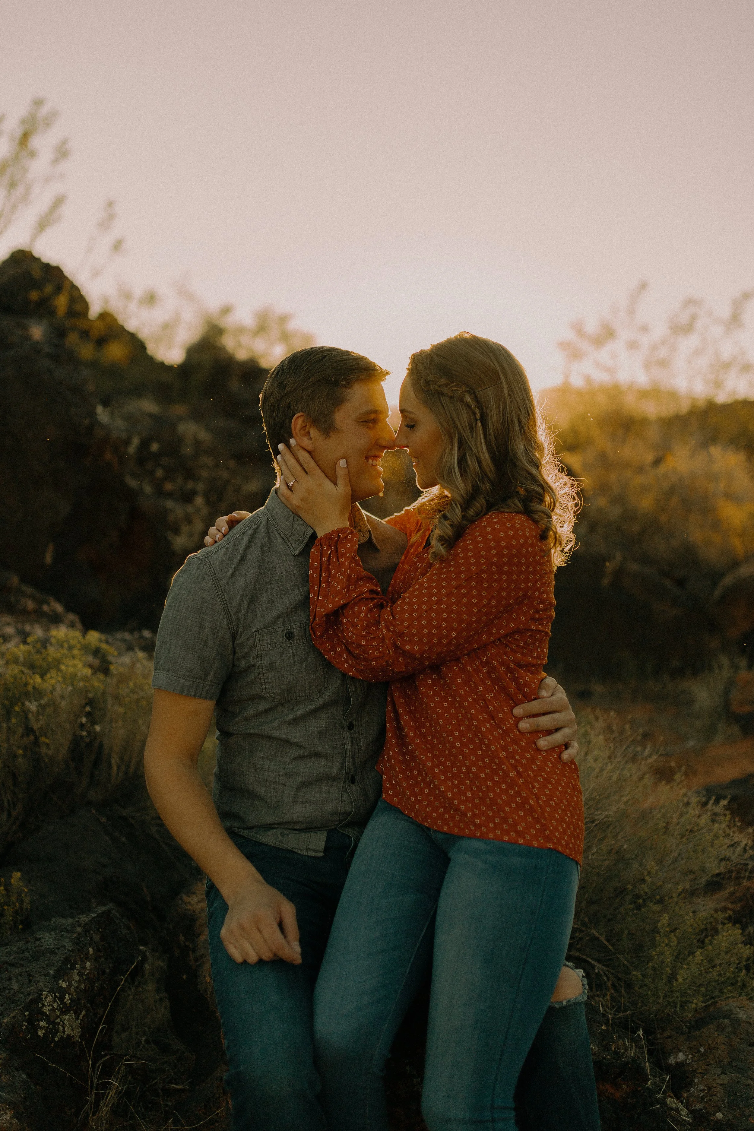 A couple sitting closely outdoors during sunset, gazing into each other's eyes, surrounded by natural landscape and rocky terrain.