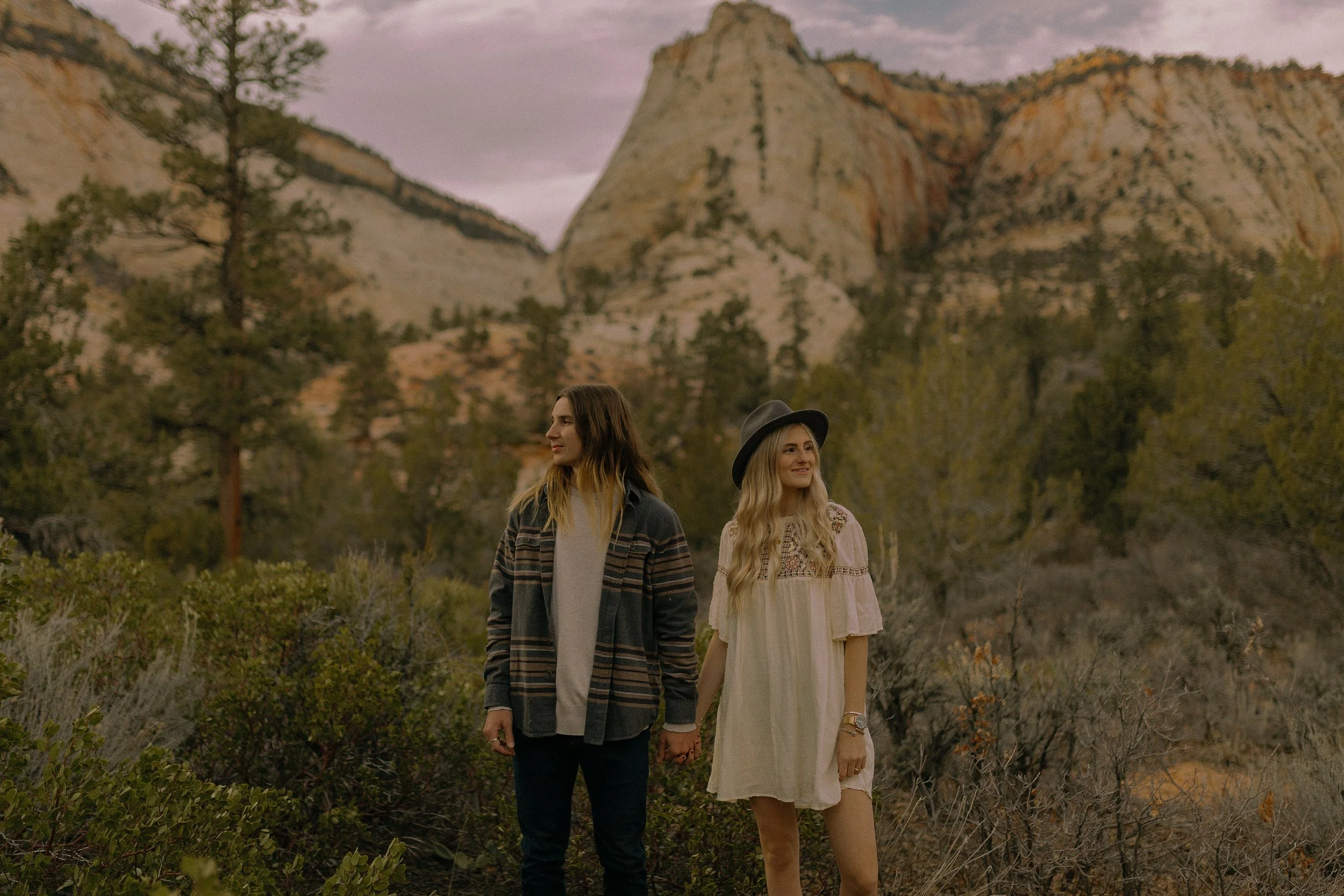 A couple standing in a scenic landscape with rocky hills and greenery, holding hands.
