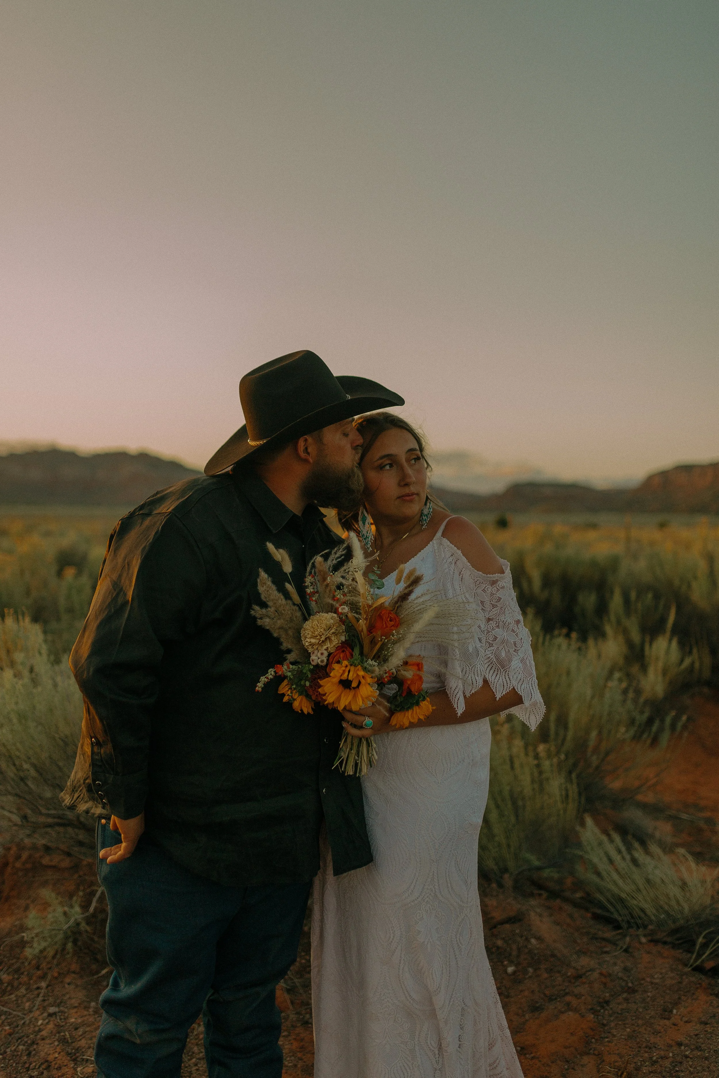 A couple standing in a scenic landscape during sunset. The man, wearing a black cowboy hat, leans in to kiss the woman, who is in a white lace dress holding a bouquet of flowers. The background features natural vegetation and mountains.