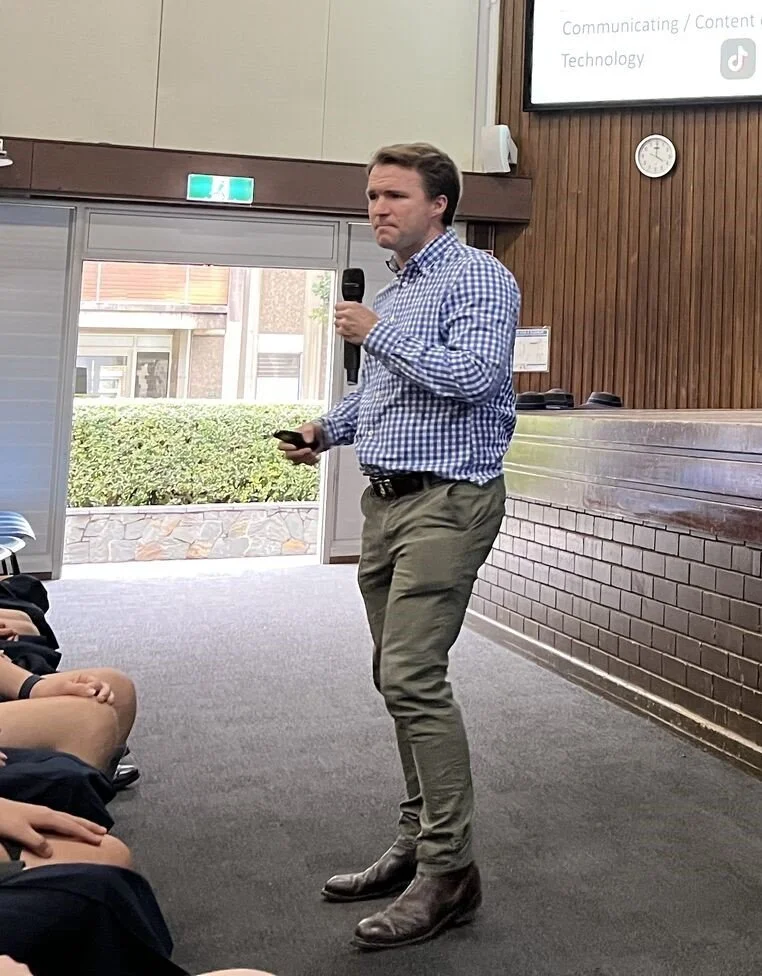 A man in a plaid shirt and khaki pants holding a microphone and a remote control stands in front of a classroom with students seated on the floor. There is a projector screen with presentation slides and a wall clock behind him.