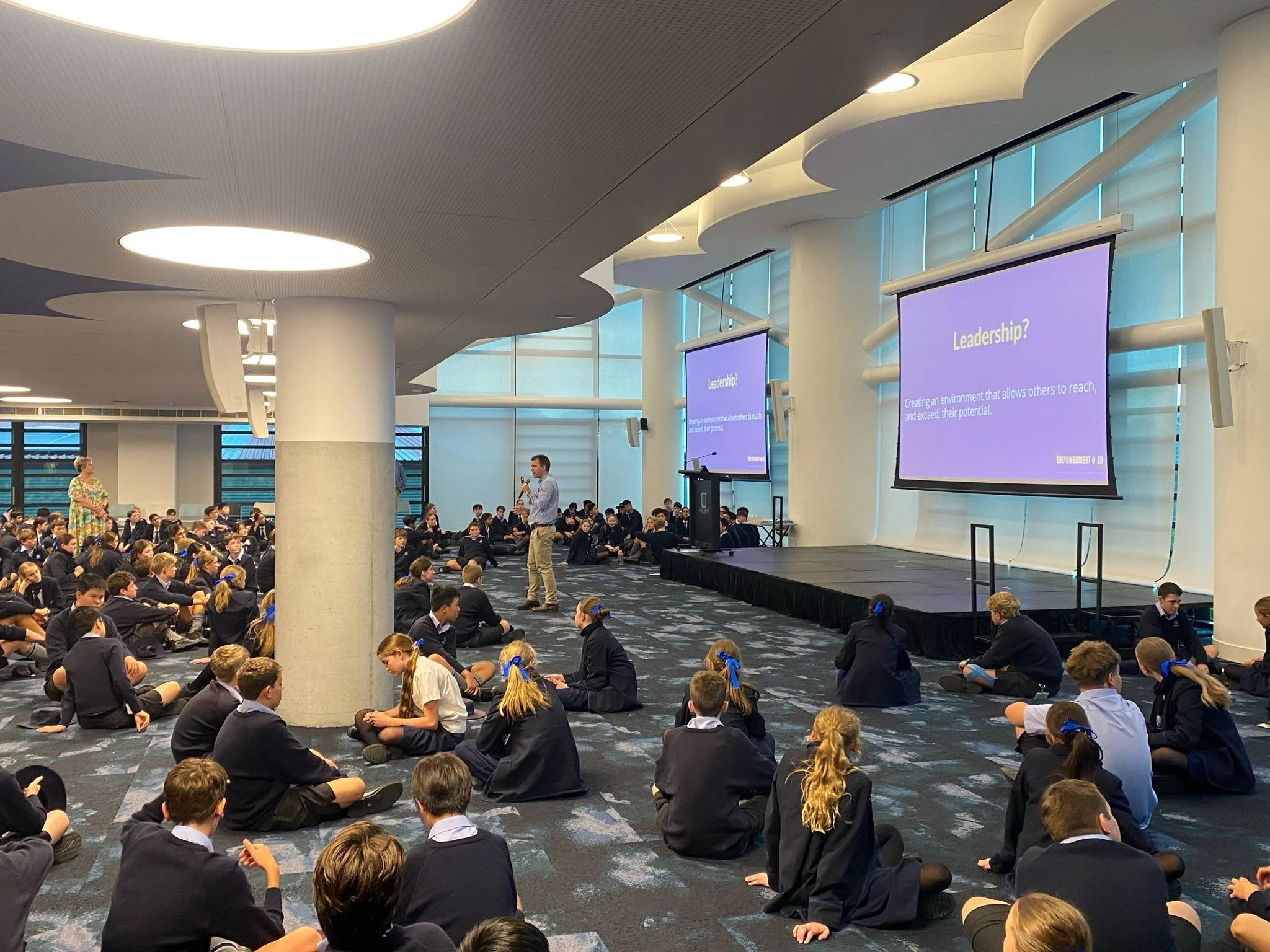 A large group of students sitting on the floor in a spacious conference room listening to a presentation. There is a stage with two large screens displaying a slide titled 'Leadership?' and a speaker holding a microphone speaking to the students.