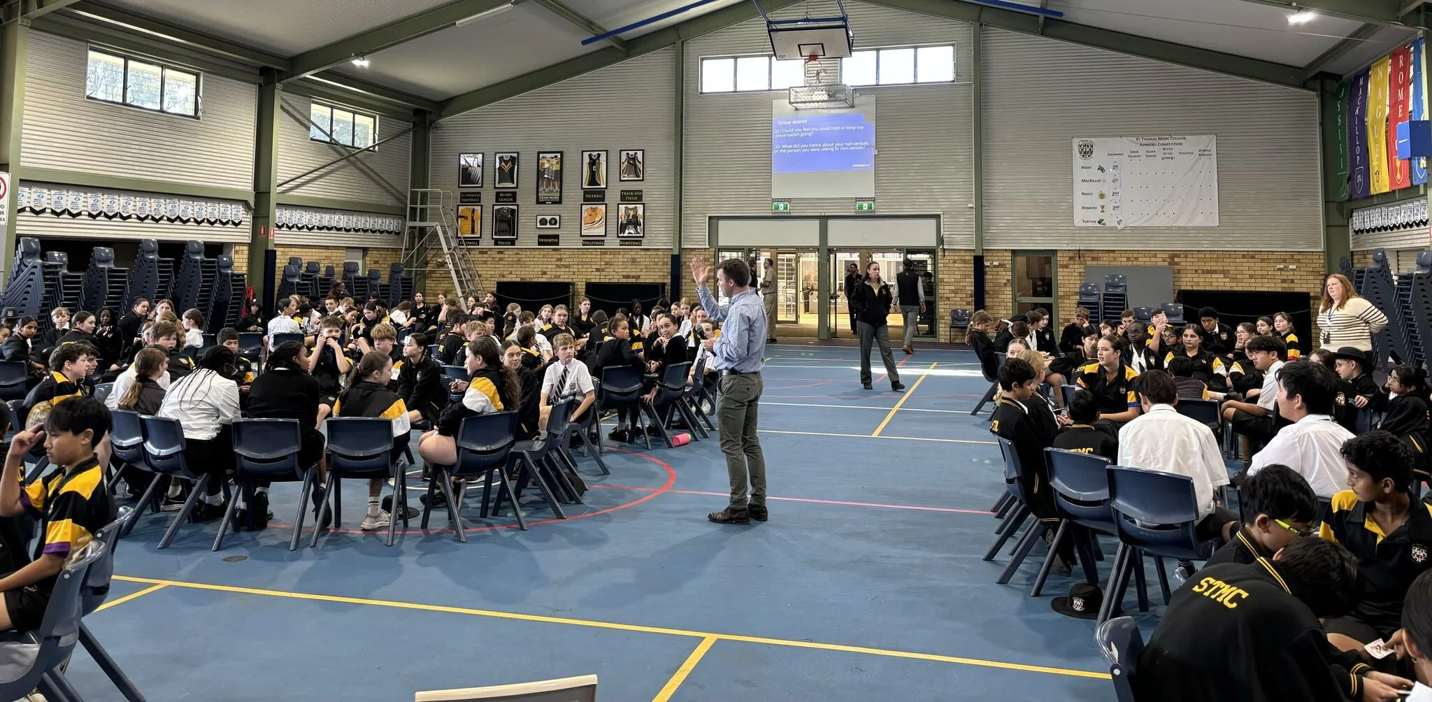 Students seated at tables in a gymnasium, with a presenter at the front. The gym has a high ceiling, windows, and banners hanging on the walls.