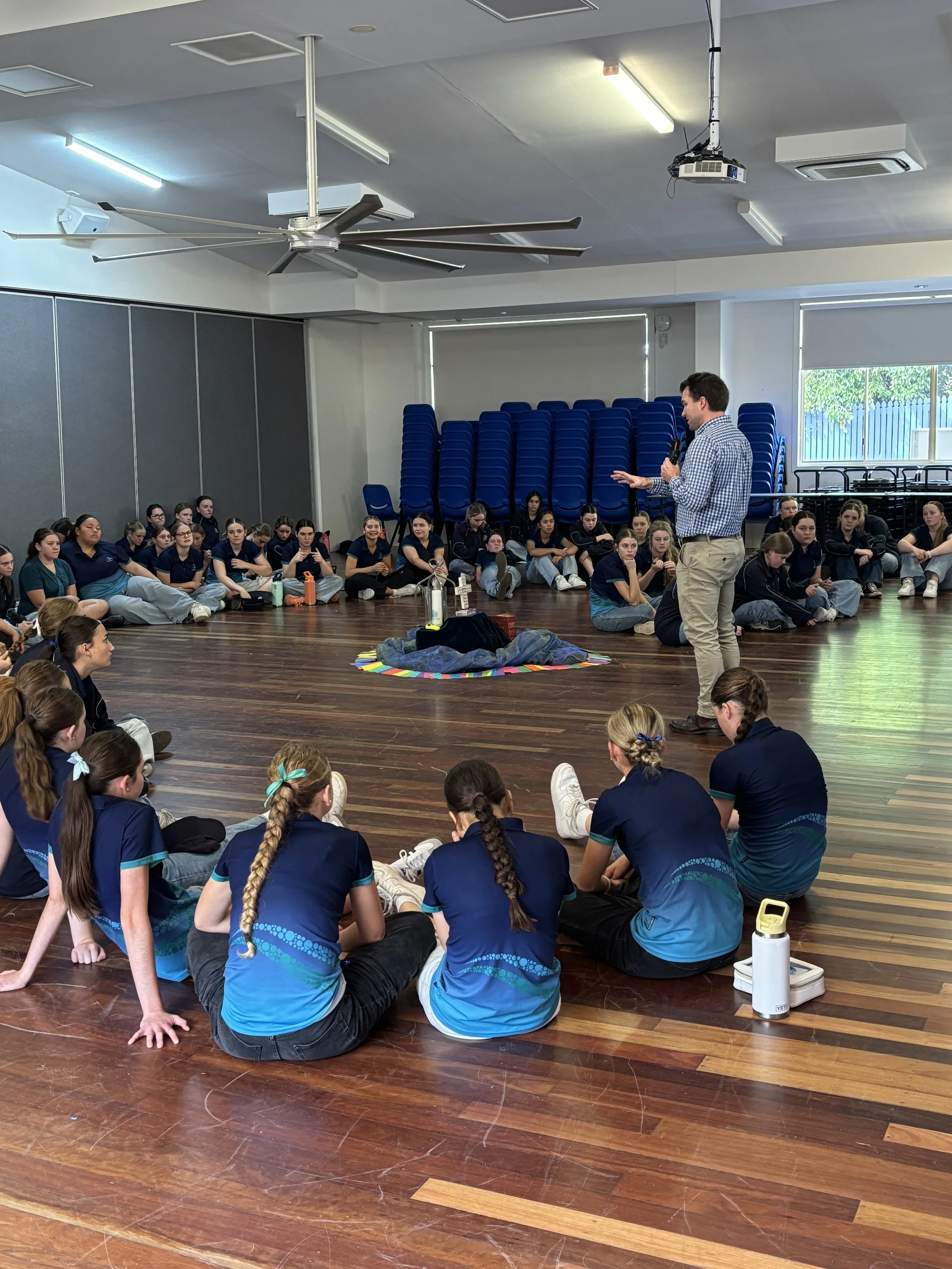 A classroom or auditorium with students sitting on the wooden floor, listening to a presenter. The presenter is a man holding a microphone, standing in front of a display area. The students are wearing dark blue and teal uniforms, and there are empty