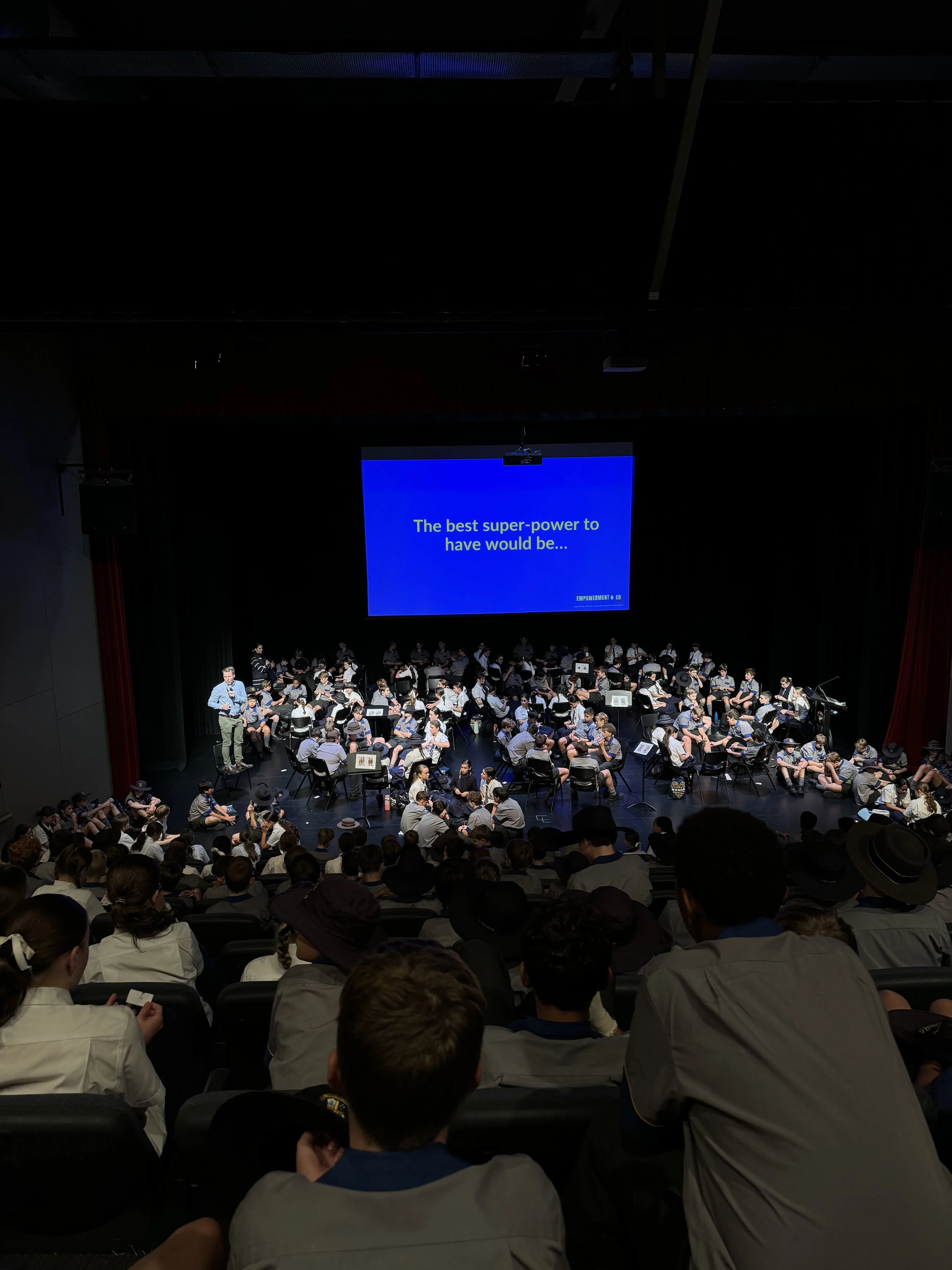 Audience seated in a theater watching a presentation with a large screen displaying text.