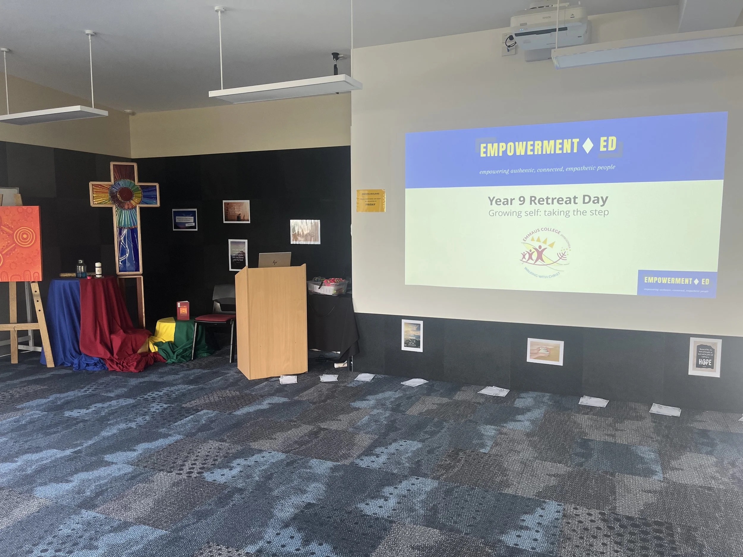 A classroom or event space set up for a retreat day with a large screen displaying a PowerPoint slide titled 'Year 9 Retreat Day,' organized by Empowerment Ed, featuring a colorful cross on a stage with fabric drapes, and framed pictures on the walls