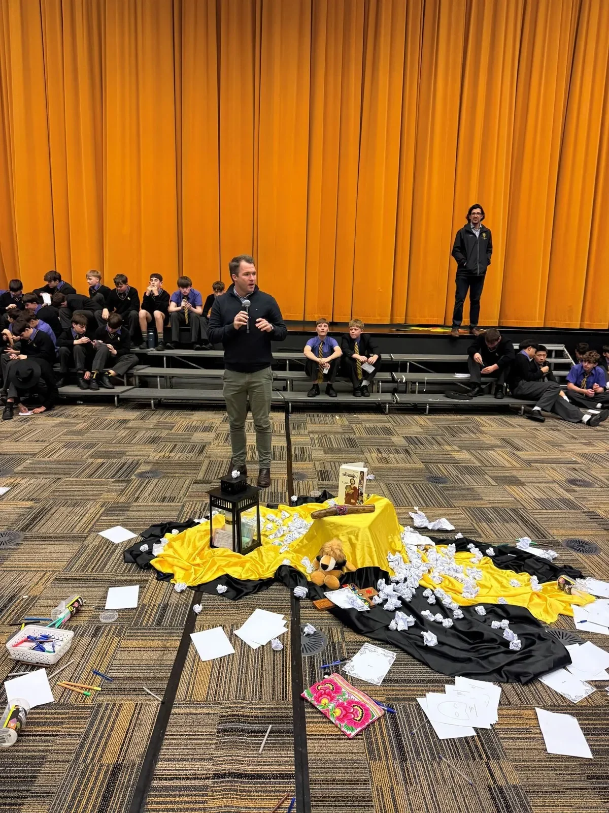 A man holding a microphone speaking in front of a group of children sitting on stairs, with a decorated table and scattered papers in front of him, in an auditorium with a large yellow curtain in the background.