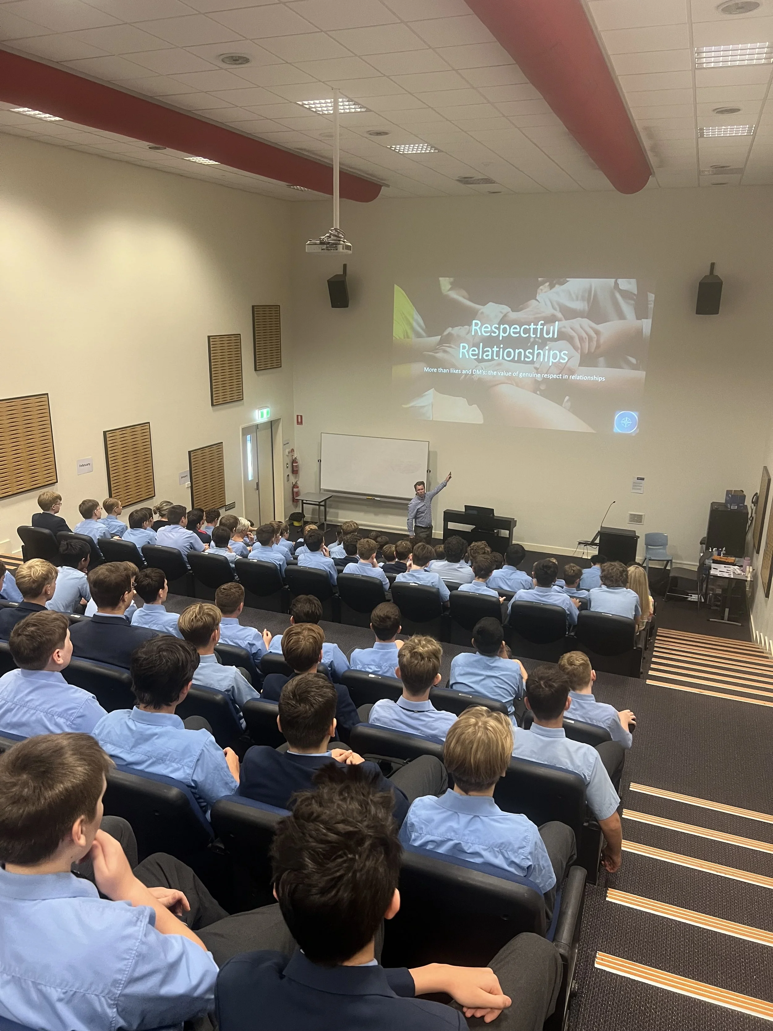 A classroom or auditorium filled with students in blue shirts attending a presentation about respectful relationships, with a speaker standing at the front pointing to a slide projected on the wall.