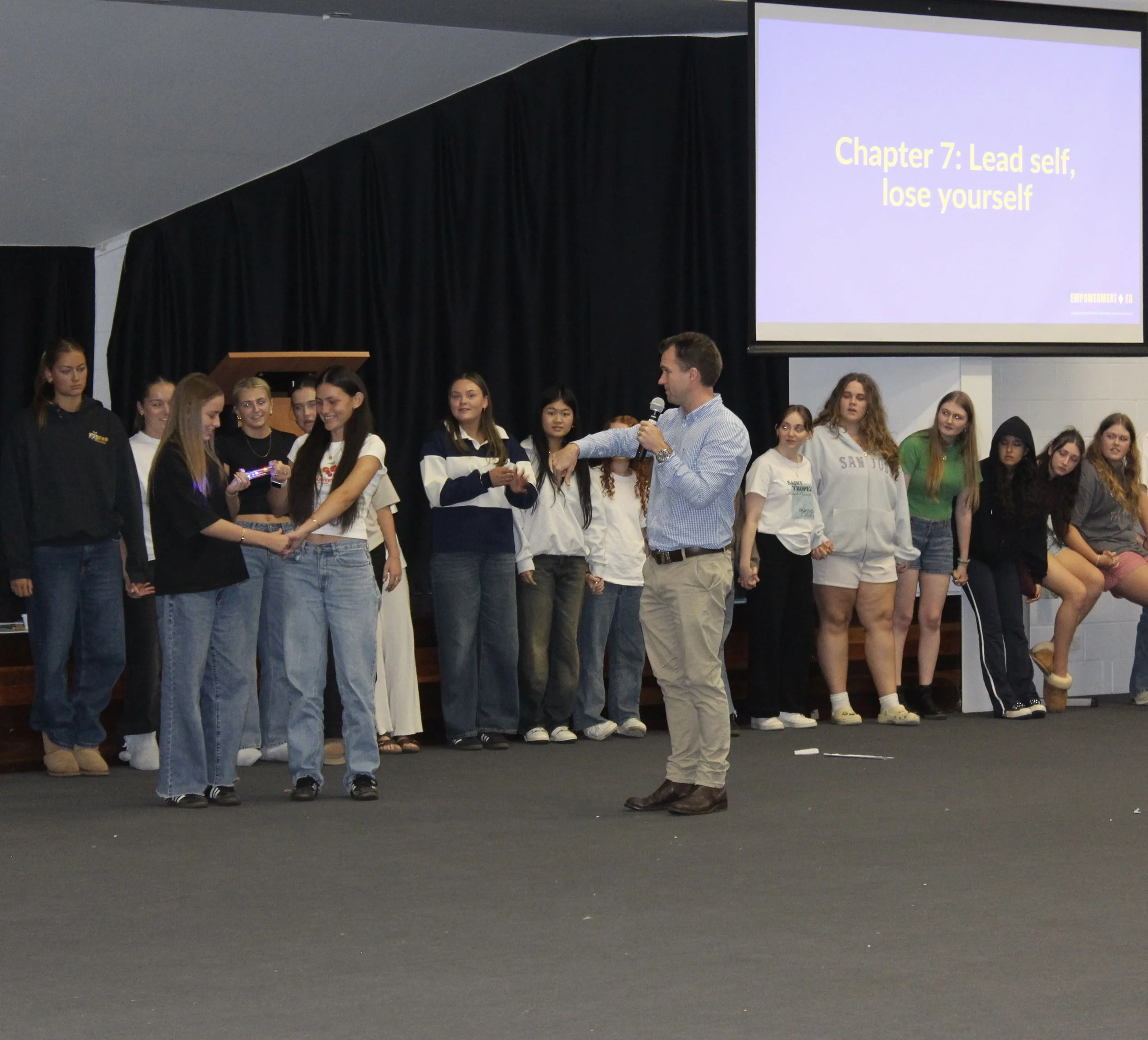 A group of young women on a stage, some holding hands, with a man speaking into a microphone. A large screen displays 'Chapter 7: Lead self, lose yourself' in yellow text.