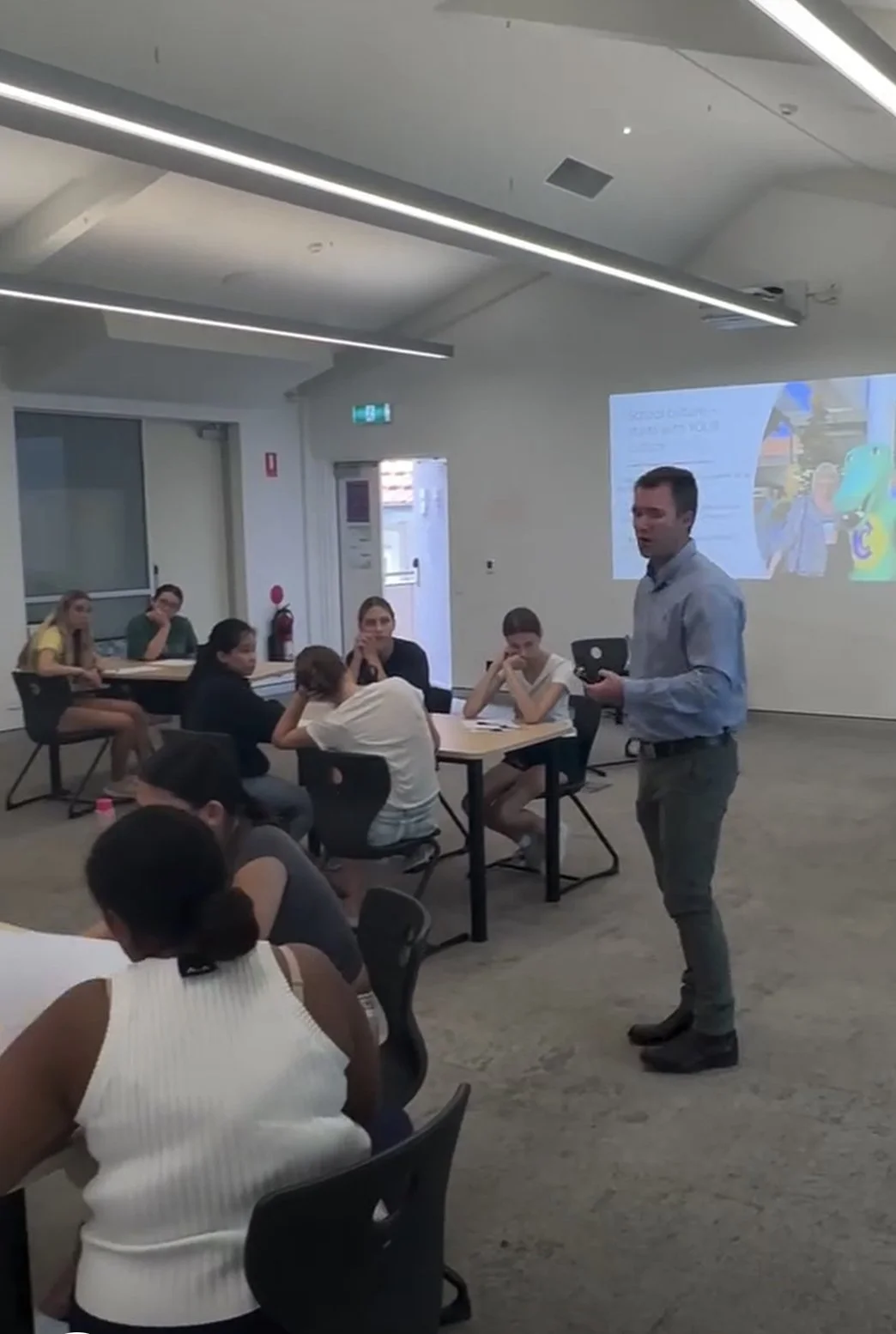 A man giving a presentation to a group of students sitting at tables in a classroom or conference room with a projection screen displaying a slide.