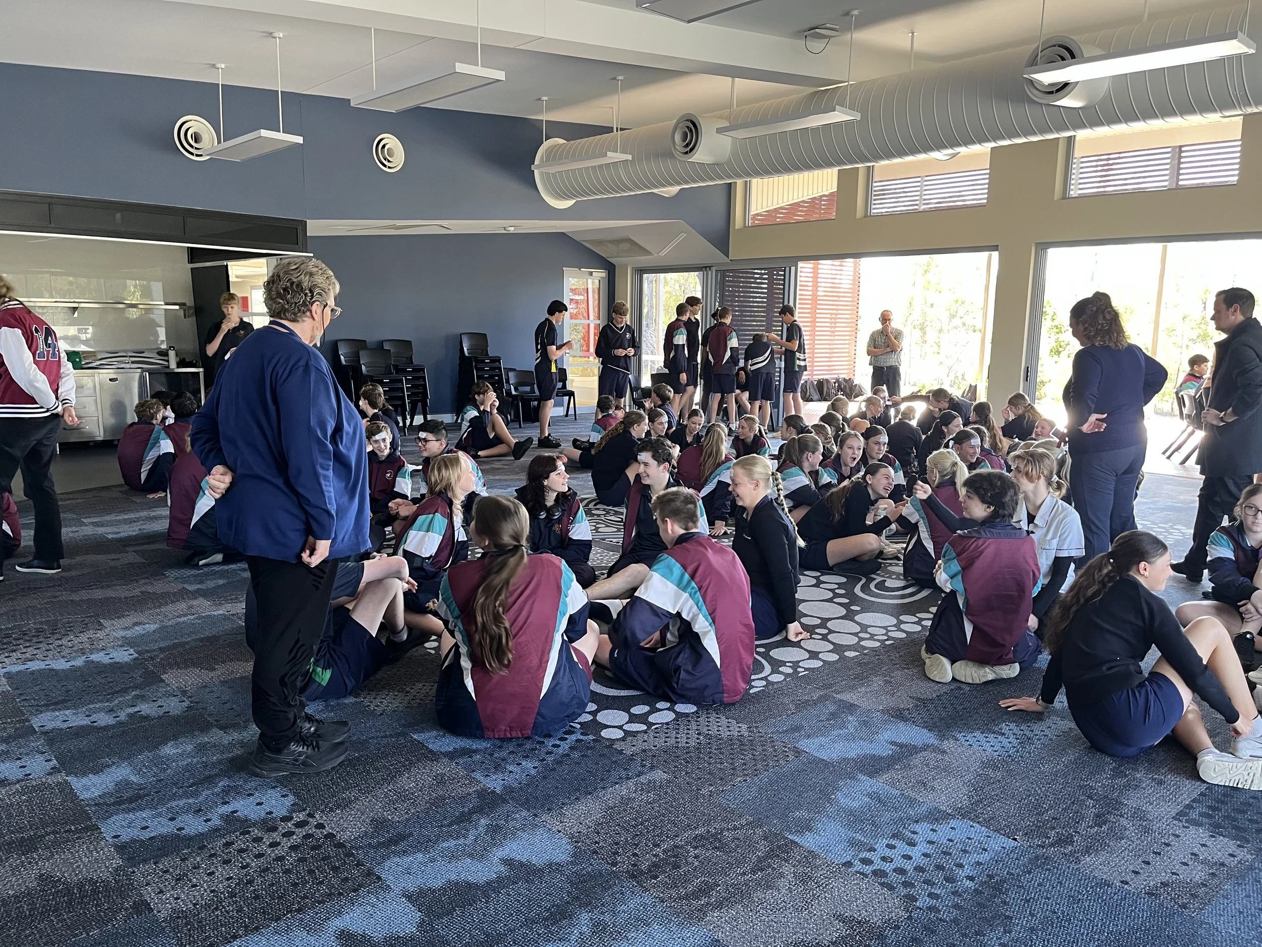 Students sitting on the floor in a large, well-lit room with large windows, while some other students and adults stand and walk around.