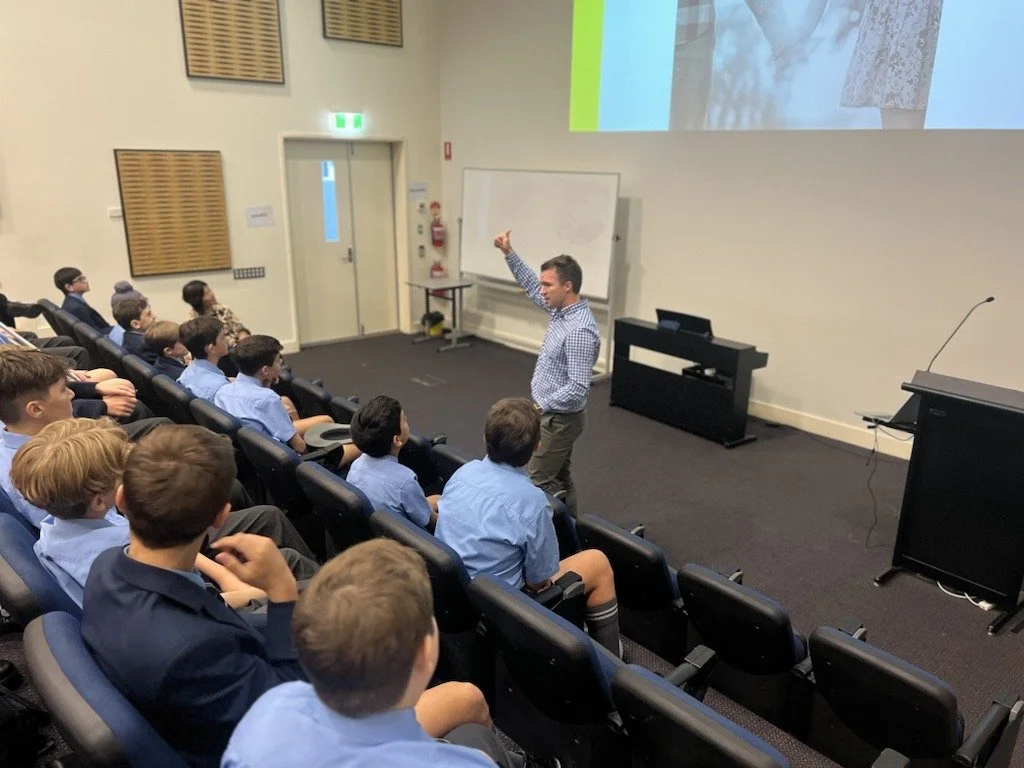 A man is giving a presentation or lecture to a group of school children seated in an auditorium. The children are wearing uniforms, some with blue shirts. The man is standing near a whiteboard, gesturing with his right hand. There is a screen above d