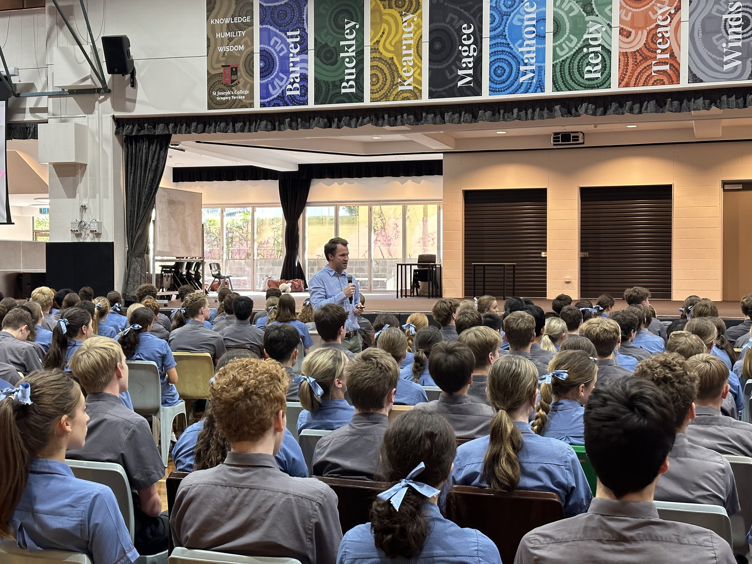 A large school assembly with students in uniform seated and listening to a speaker on stage inside a school auditorium.
