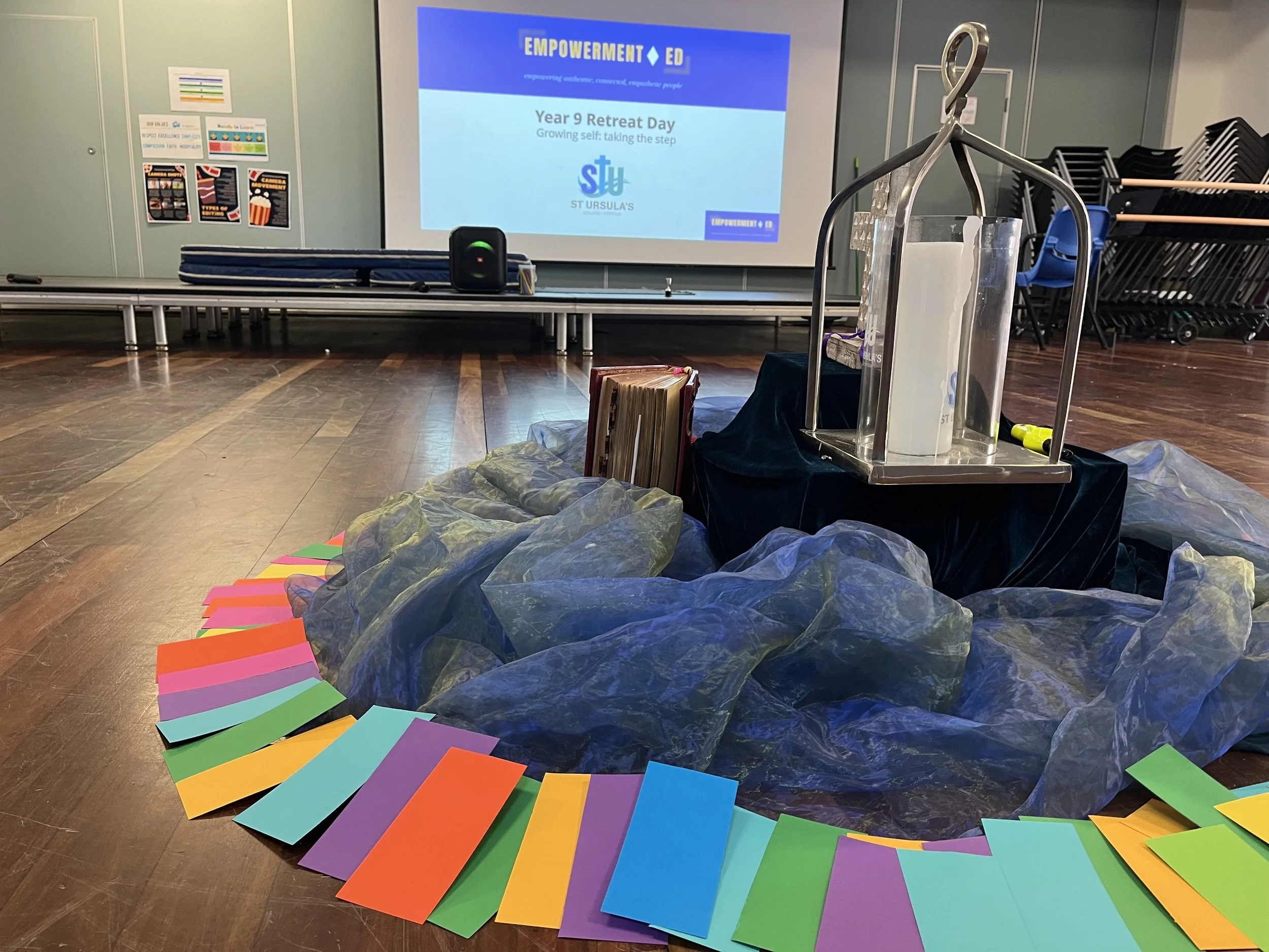 Decorative display with water jug and books on a black cloth, surrounded by colorful paper, in front of a presentation screen with the title 'Year 9 Retreat Day' and the logo of ST Ursula's.