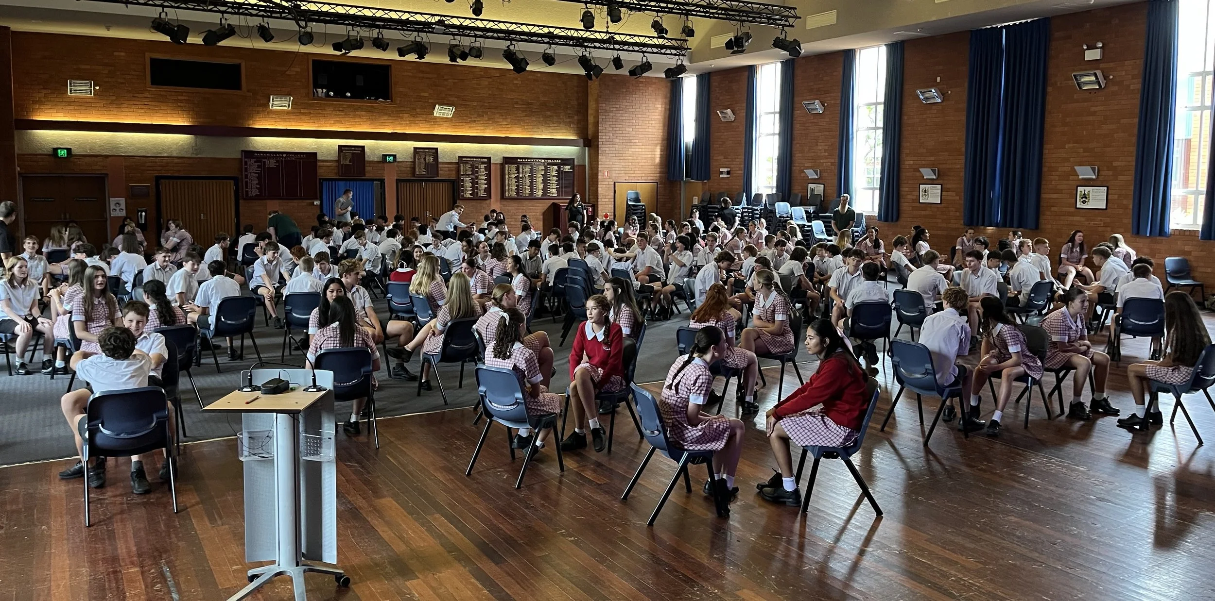 A large indoor hall filled with students in school uniforms sitting in chairs, some engaging in conversations, with a few standing or walking. The room has brick walls, large windows with blue curtains, and a wooden floor. There is a podium with a mi