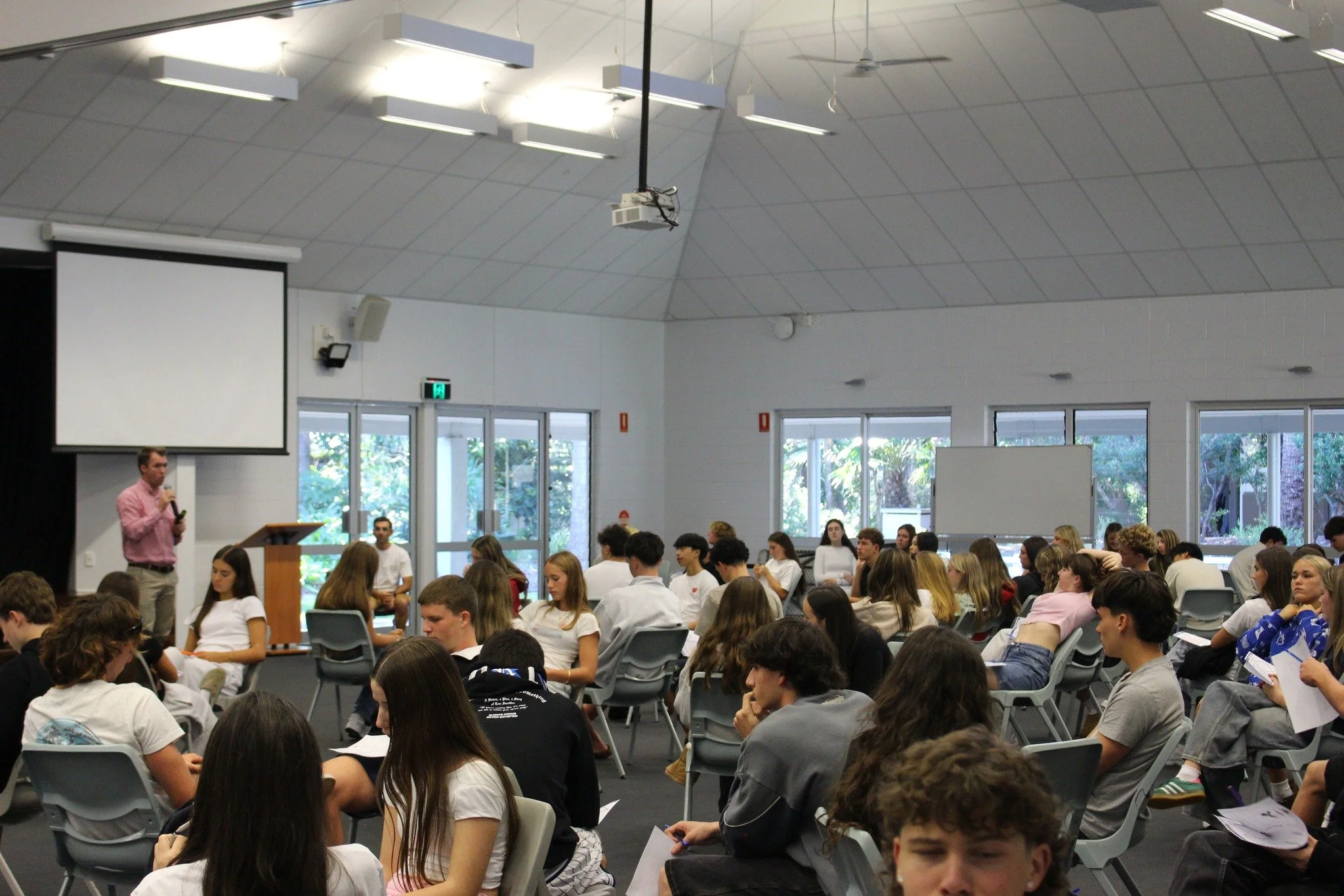 A large group of students seated in a classroom or auditorium listening to a speaker standing at a podium. The room has a high, sloped ceiling with lights and large windows in the background.