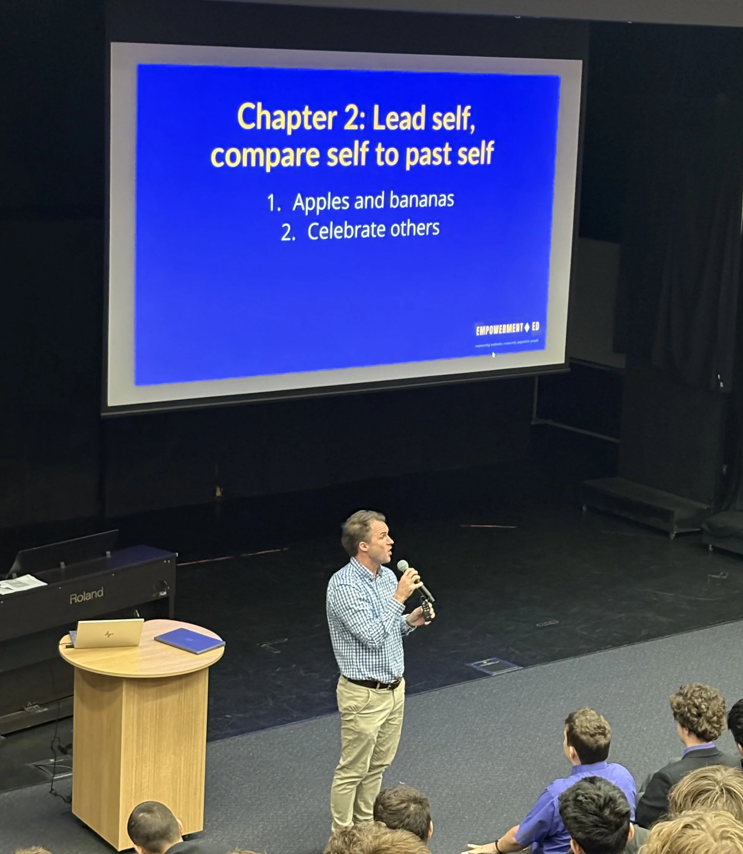 A man holding a microphone giving a presentation to an audience. The slide on the screen behind him reads 'Chapter 2: Lead self, compare self to past self' with two bullet points: 1. Apples and bananas, 2. Celebrate others. The setting appears to be