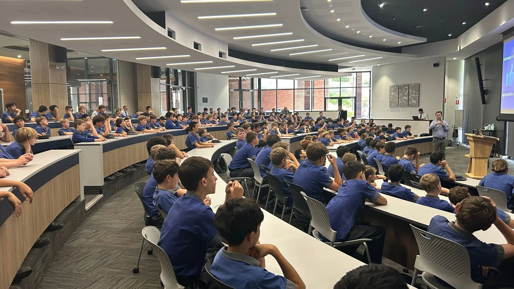 A large classroom with many students in blue uniforms seated at desks, facing a presenter and a large screen, during a lecture or presentation.