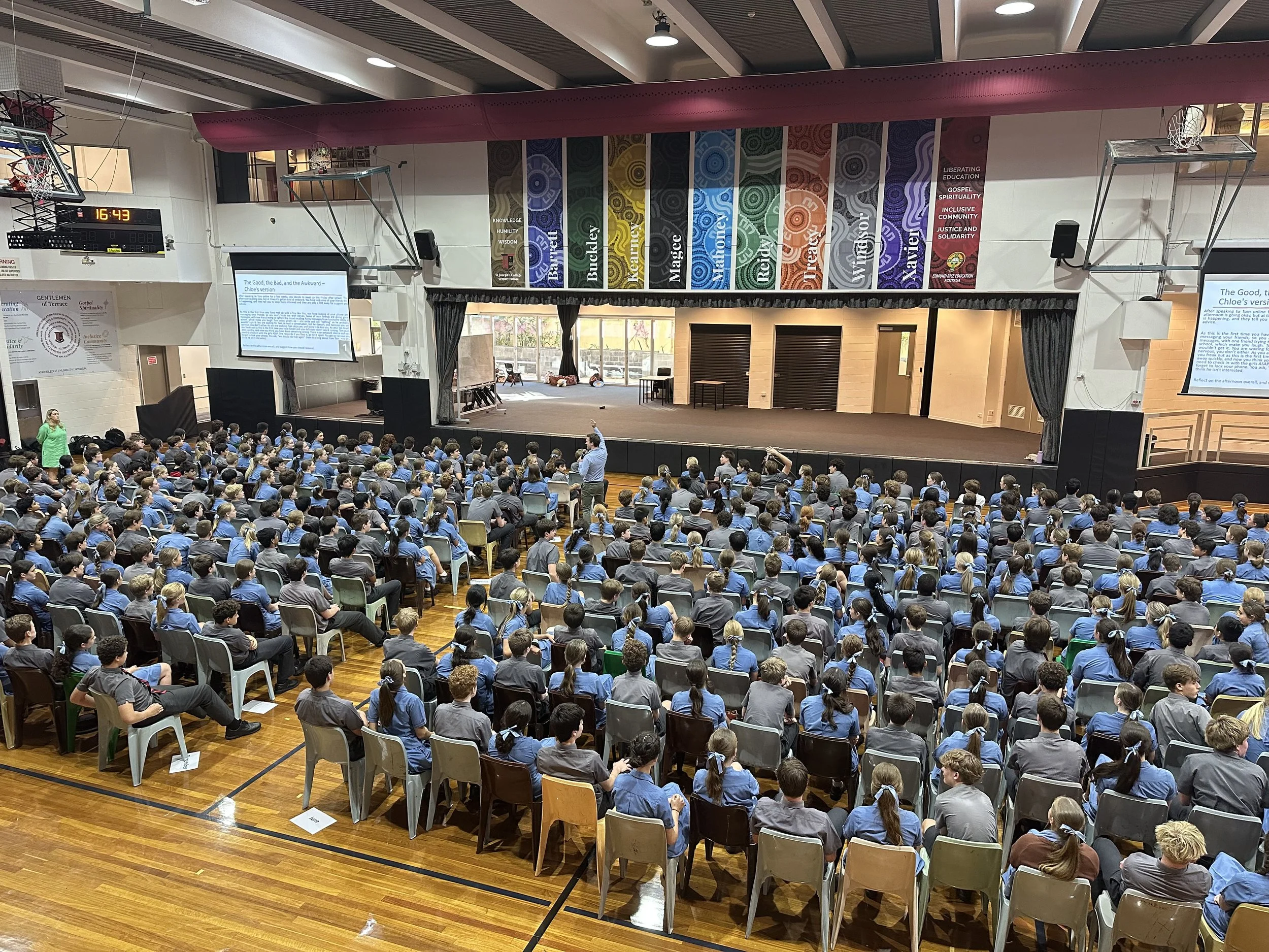 Large indoor auditorium filled with students sitting in chairs, facing a stage with two large screens showing a presentation, and colorful banners hanging above.