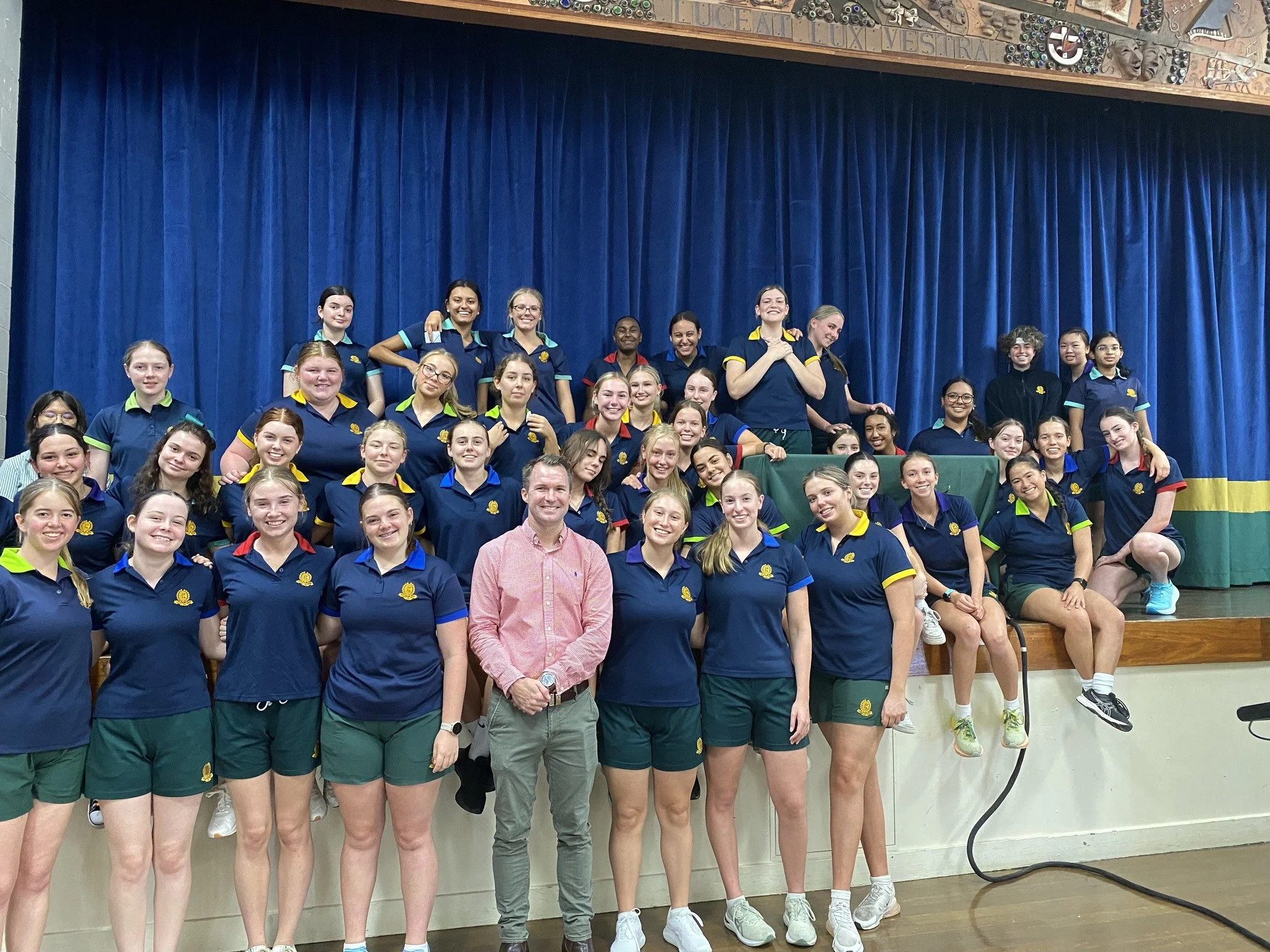 Group of young women in navy athletic shirts and green shorts on stage with a man in a pink shirt, in front of blue curtain, smiling for a group photo.