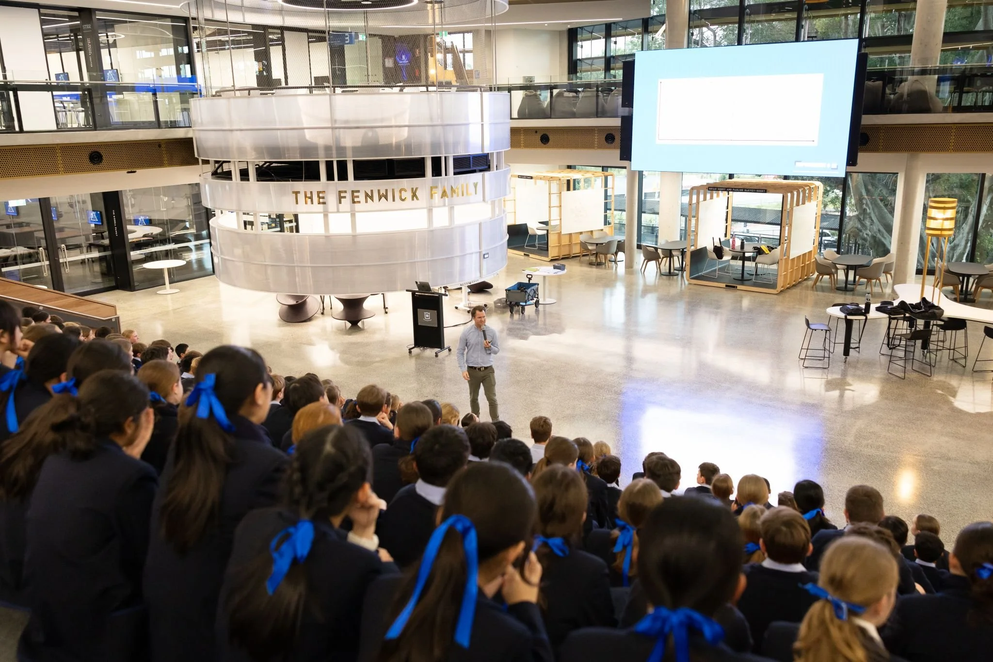 A man giving a presentation to a large group of students in a modern indoor space with glass walls and seating areas.