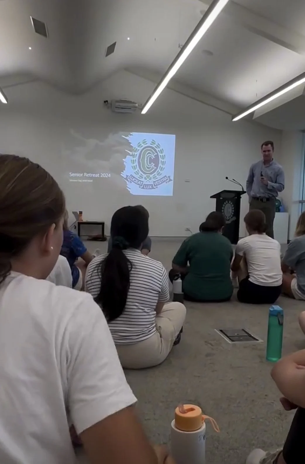 A group of students seated on the floor of a classroom or seminar room, listening to a man giving a presentation. A projected slide displays the logo for 'Senior Retreat 2024' with some text underneath.