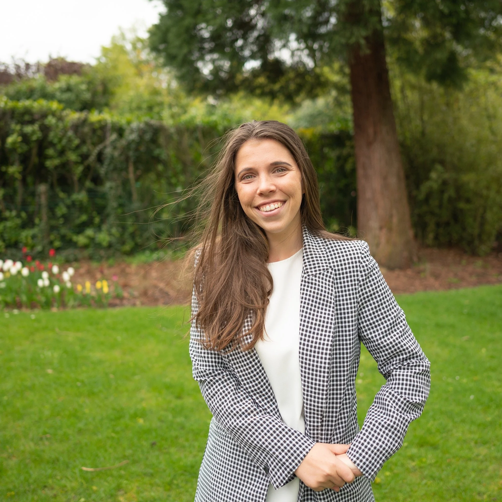 A woman standing outdoors in a park with trees and grass, smiling at the camera.