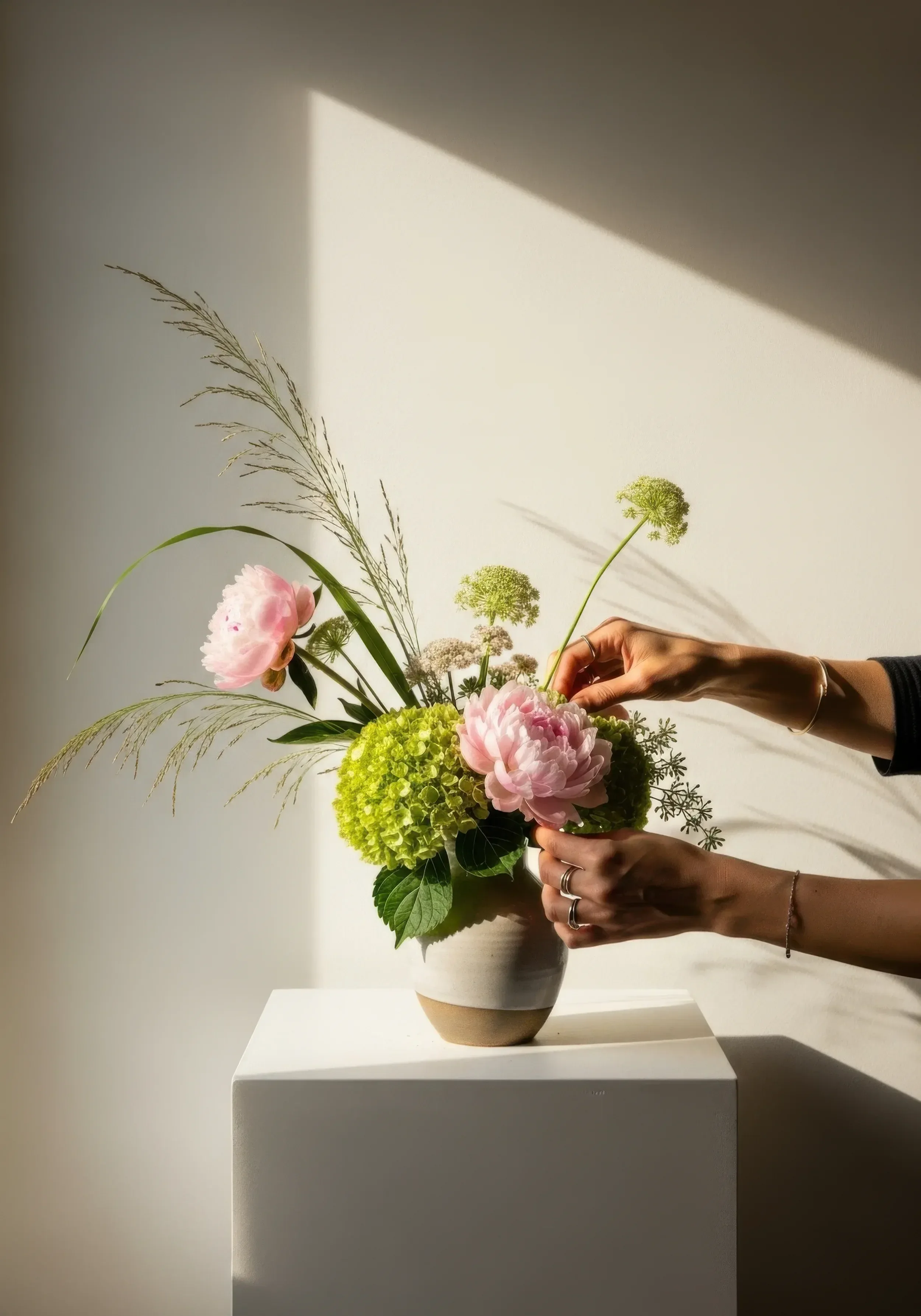 Bukiet kwiatów w beżowym wazonie, kwiaty to różowa piwonia, zielone hortensje i zielone trawy, ręce dotykają kwiatów.