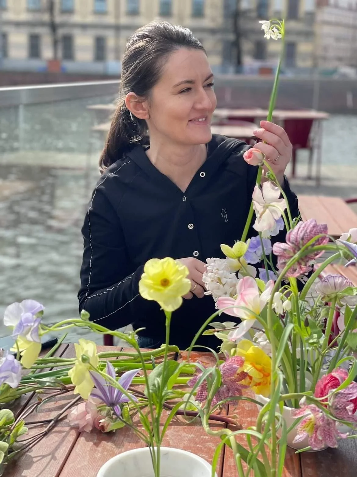 Kobieta o ciemnych włosach siedzi przy stole, uśmiecha się i trzyma kwiat w dłoniach, na stole przed nią leżą kwiaty i stoją białe miseczki z kwiatami.