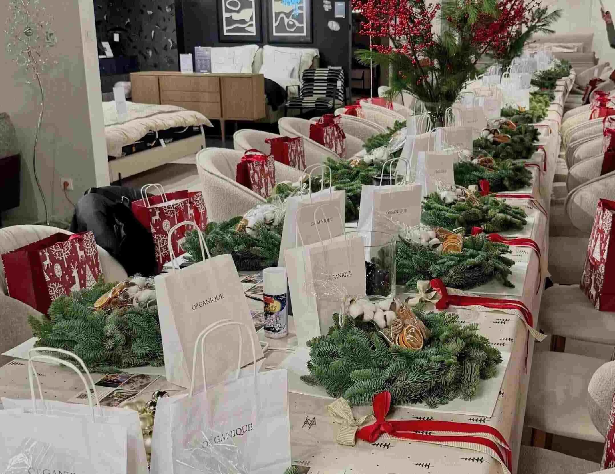 Stół przygotowany do warsztatów, na stole znajdują się zielone wianki, naturalne dekoracje, wstązki, wazony z zielonymi gałęziami sosny i gałęzie ileksu, sztuczny śnieg i torby papierowe z opominkiem.