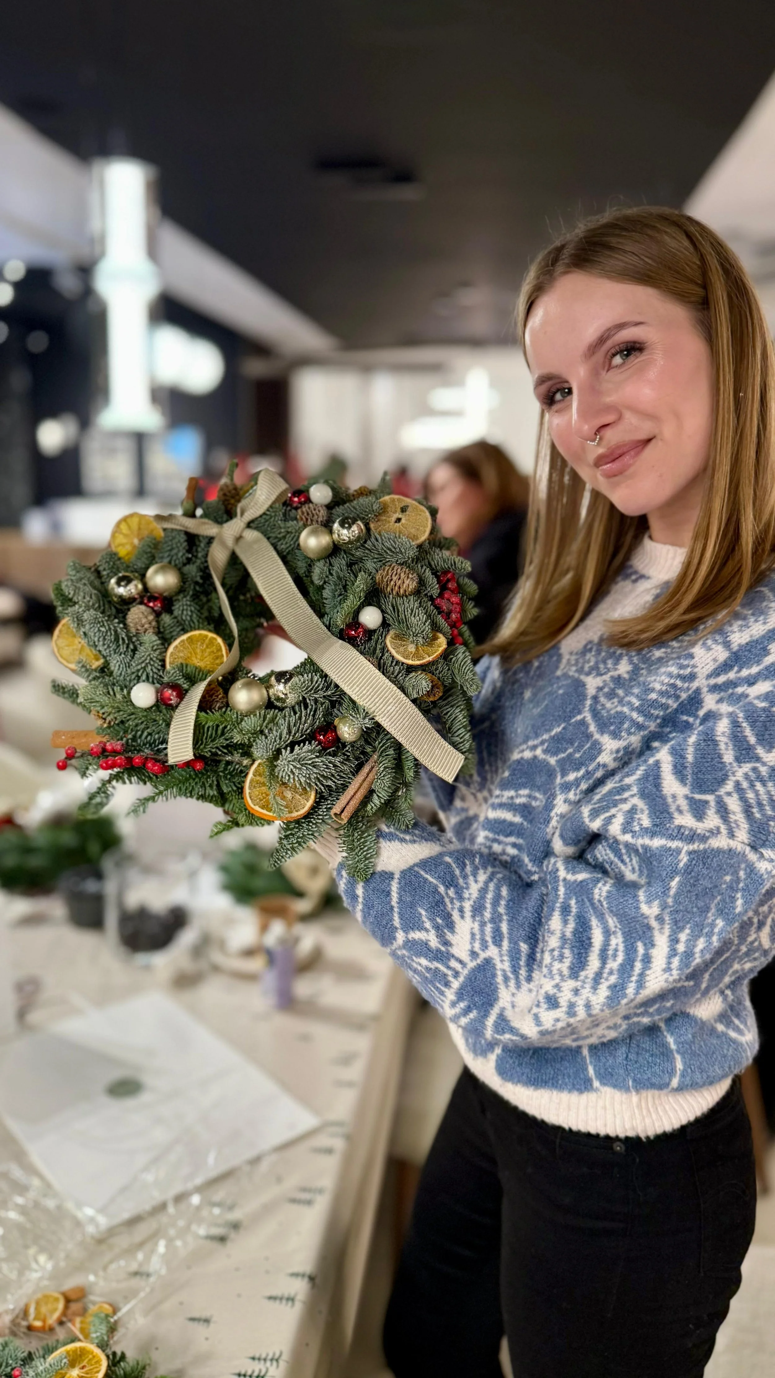 Kobieta z długimi rudymi włosami, ubrana w niebiesko-biały sweter, trzyma w pokoju ozdobny wieniec świąteczny z plastrami pomarańczy, szyszkami i bombkami.