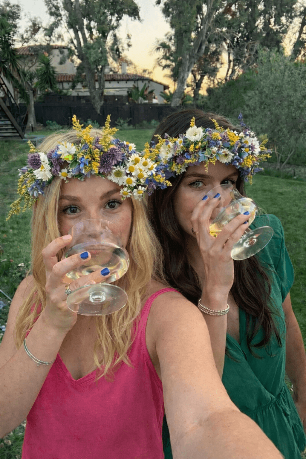 Dwie kobiety w różowej sukience i zielonej z wiankami na głowach piją z kieliszków prosecco w plenerze.