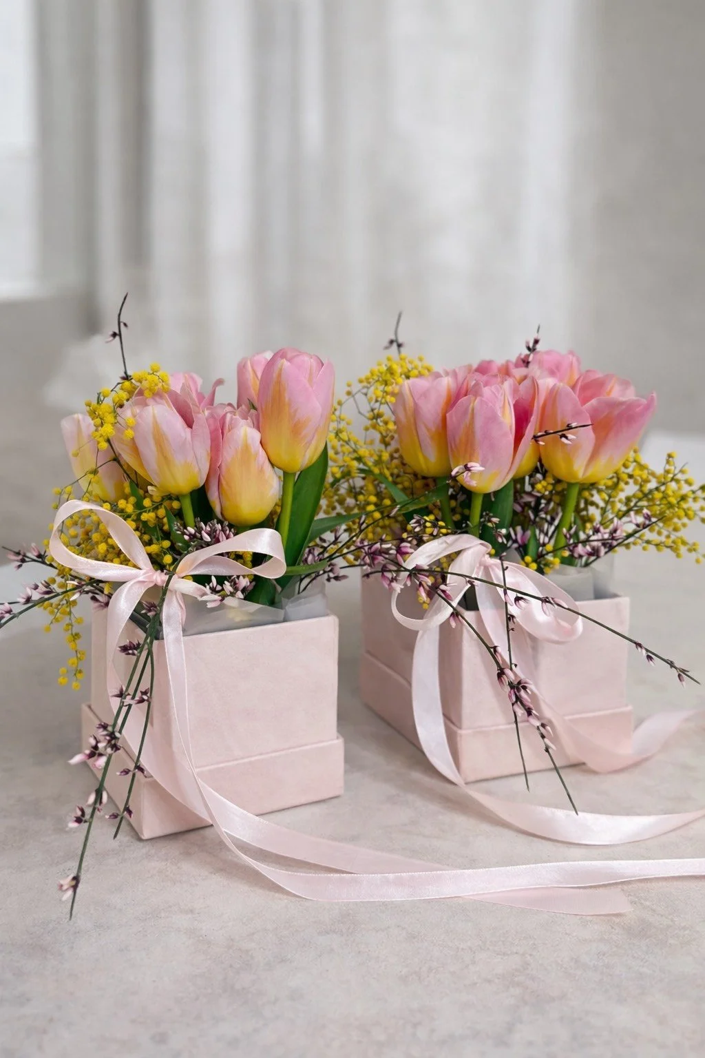 Dwa  kwiatowe flowerboxy z żółtymi tulipanami i żółtymi kwiatami w jasnoróżowych kwadratowych pudełkach, z różowymi wstążkami, na białej powierzchni.