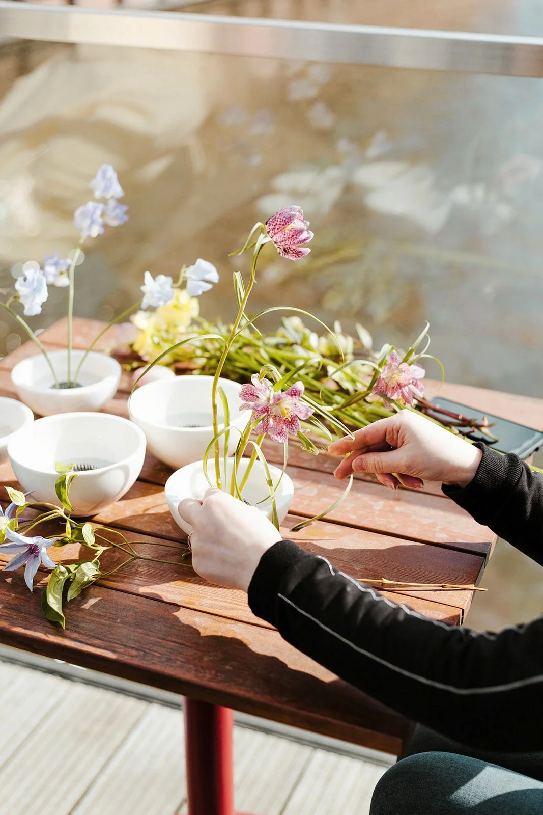 Osoba układająca kwiaty w białych misach na drewnianym stole, wokół którego porozrzucane są różne kwiaty i liście.