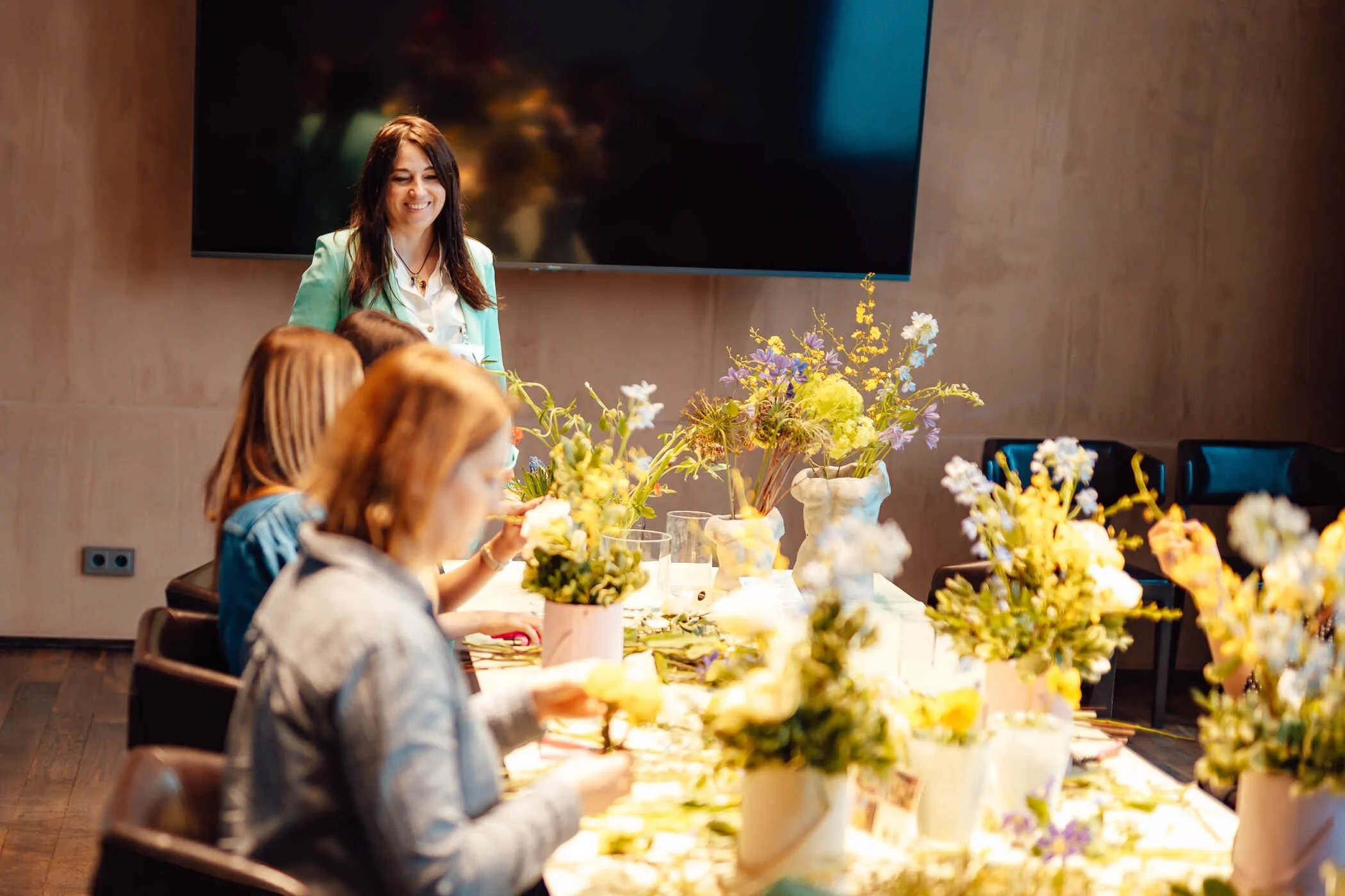 Kobiety siedzą przy długim stole, na którym są kwiaty i układają kwiatowe flowerboxy, a prowadząca stoi obok stołu w zielonym garniturze i się uśmiecha do nich.